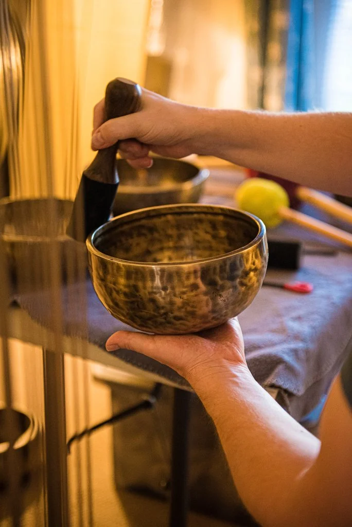 Person holding a singing bowl with a wooden mallet in a peaceful setting with musical instruments in the background.