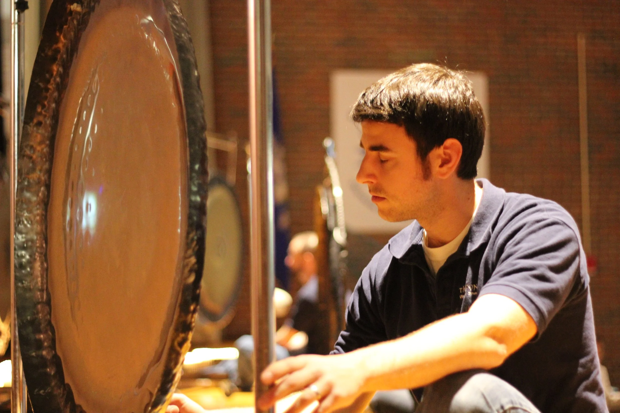 Young man playing a gong in an indoor setting with a focused expression.