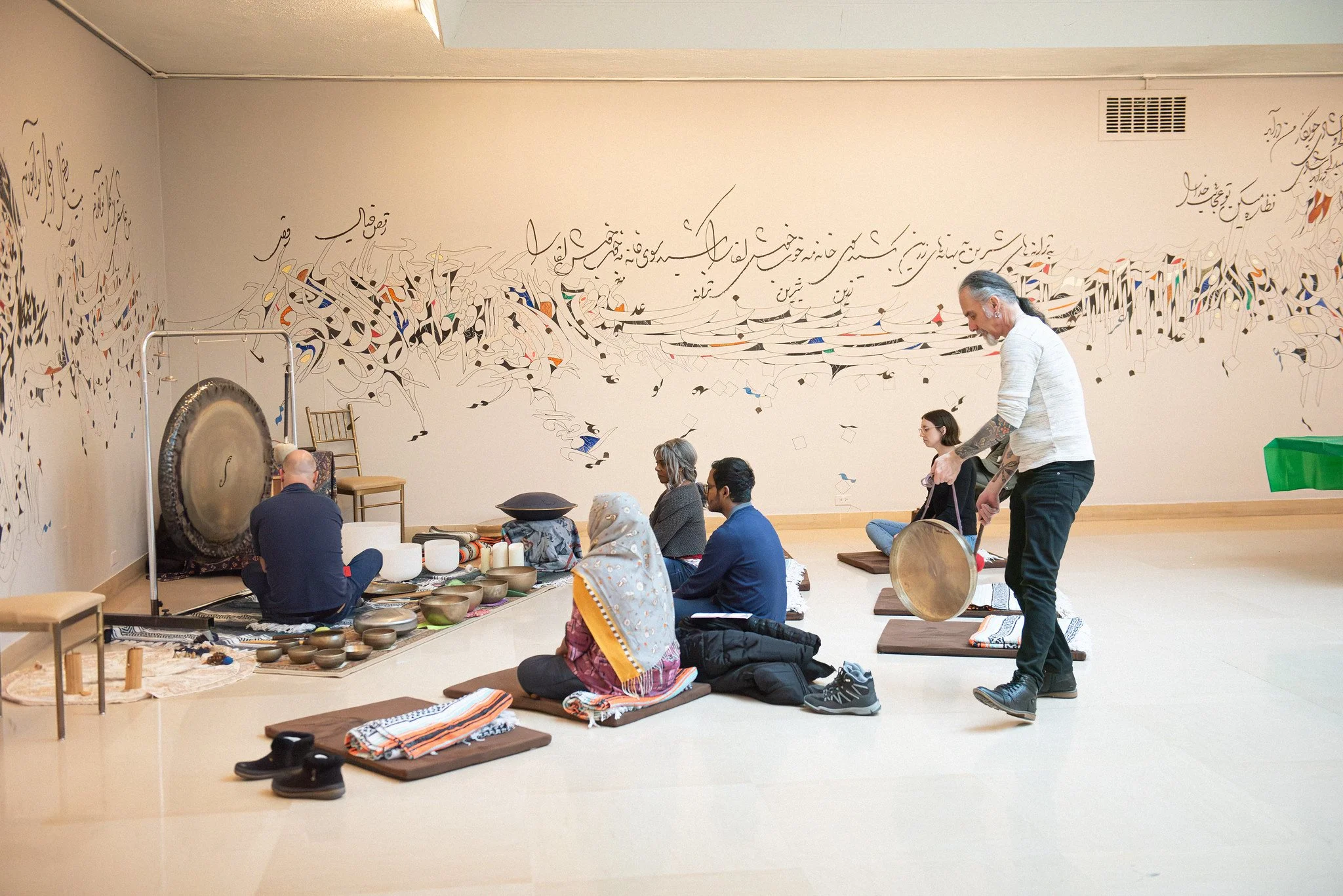People participating in a sound bath meditation in a room with a large mural of calligraphy on the wall. They are seated on cushions and mats, with a person playing a gong and other instruments.