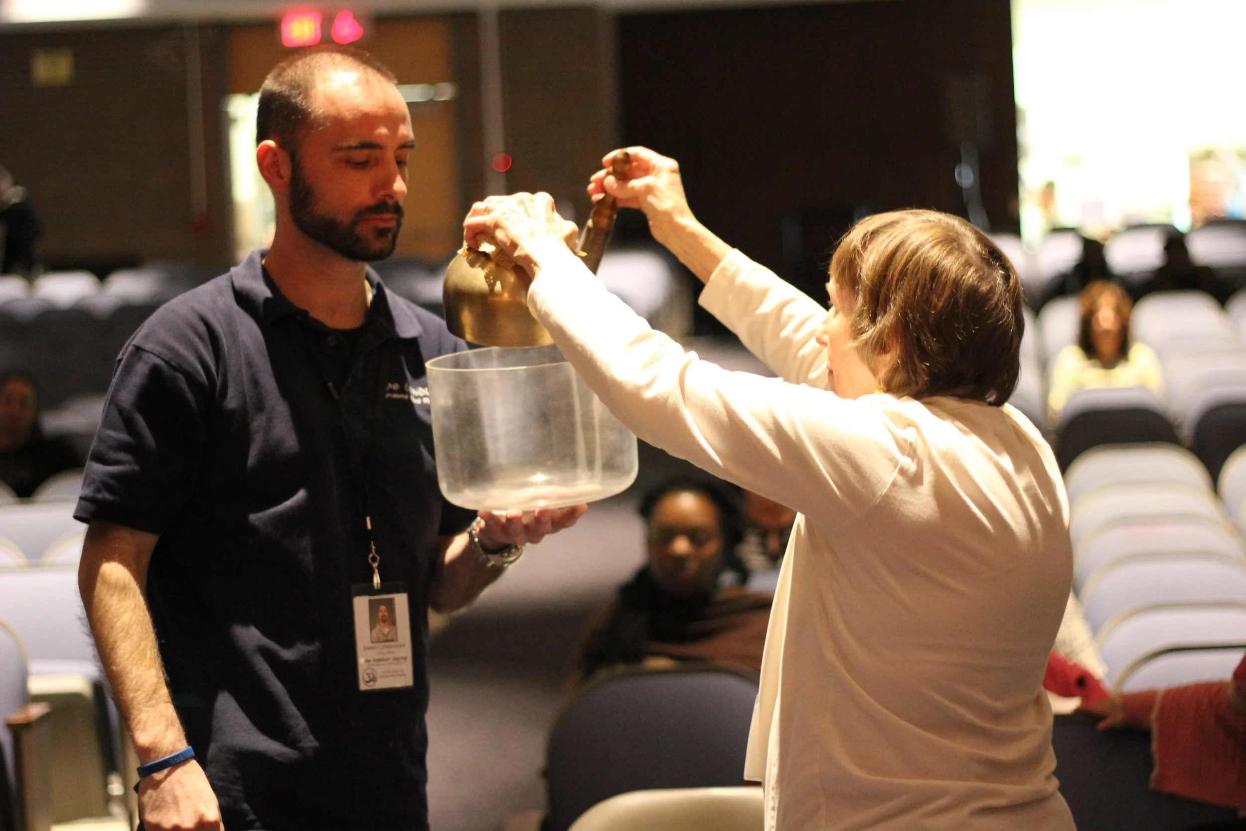 A woman and a man participating in a ceremonial activity on stage. The woman is holding a brass bell and a glass container, while the man is holding a large glass bowl. The setting appears to be a conference or seminar with an audience seated in rows