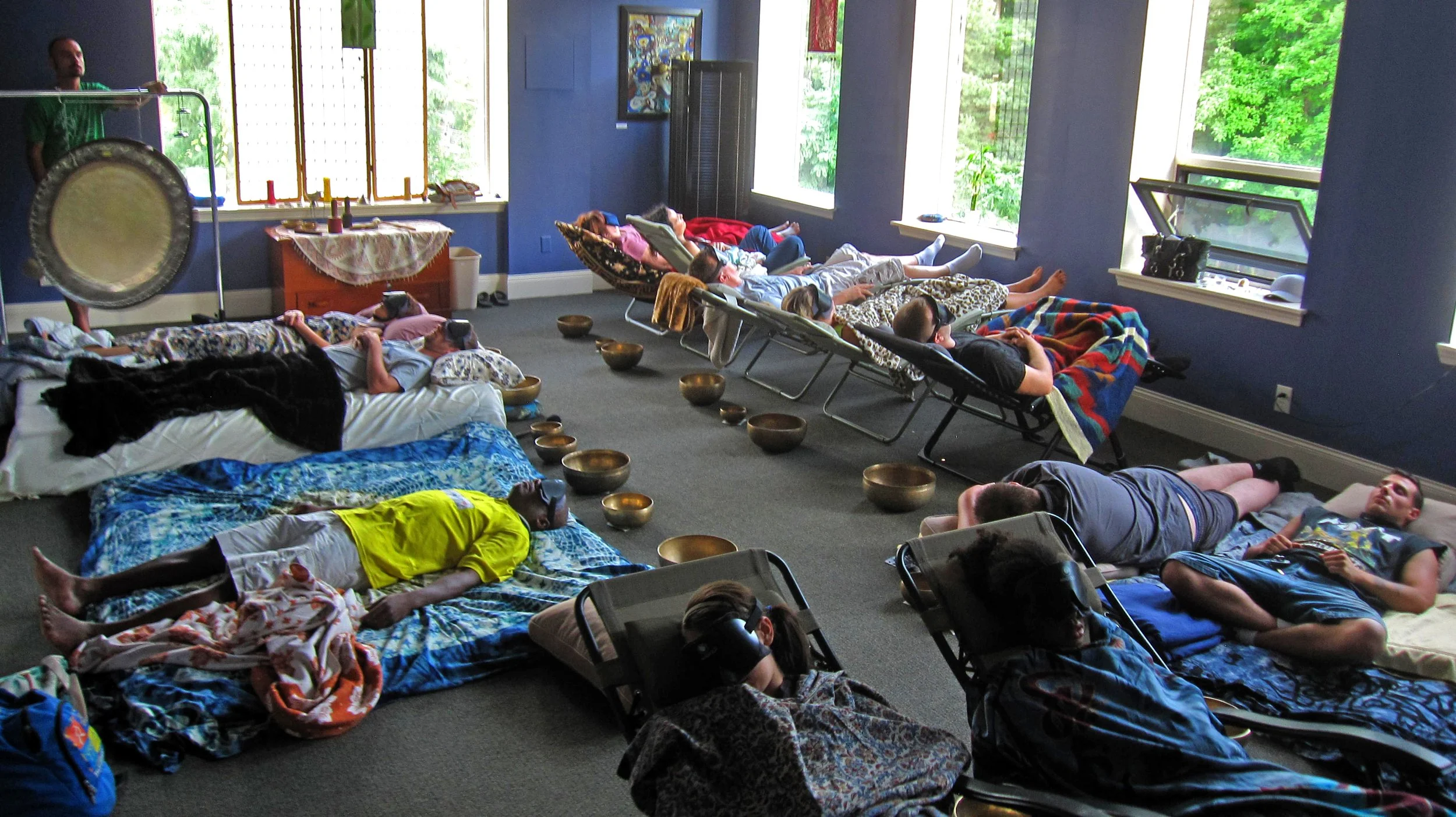 People lying on beds and lounge chairs, listening to a person playing gong, in a room with large windows and artwork on walls.