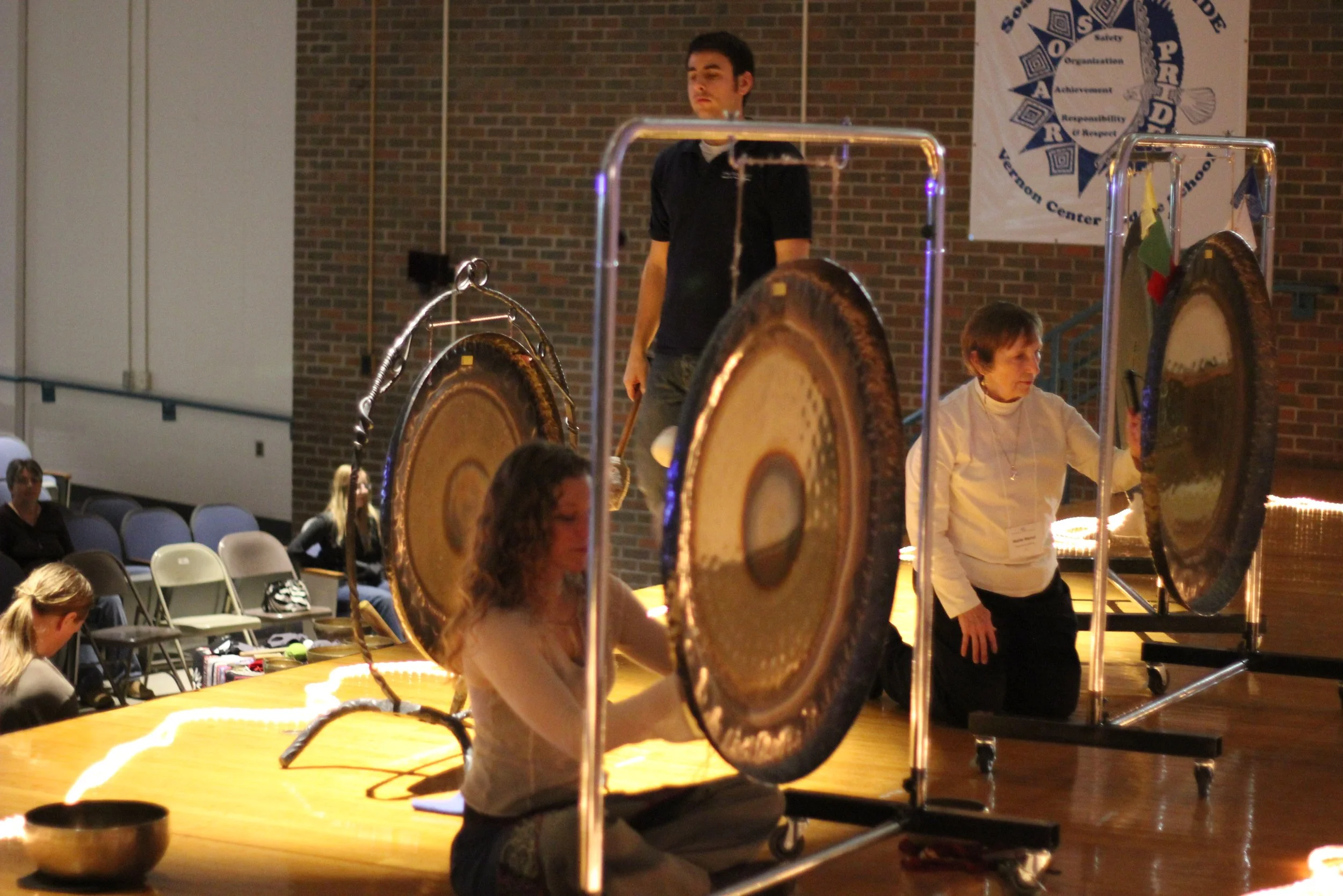 Three people performing on stage with large gongs, one woman sitting and striking a gong, another woman kneeling beside her, and a man standing in the background, in an indoor auditorium with a brick wall and a banner.