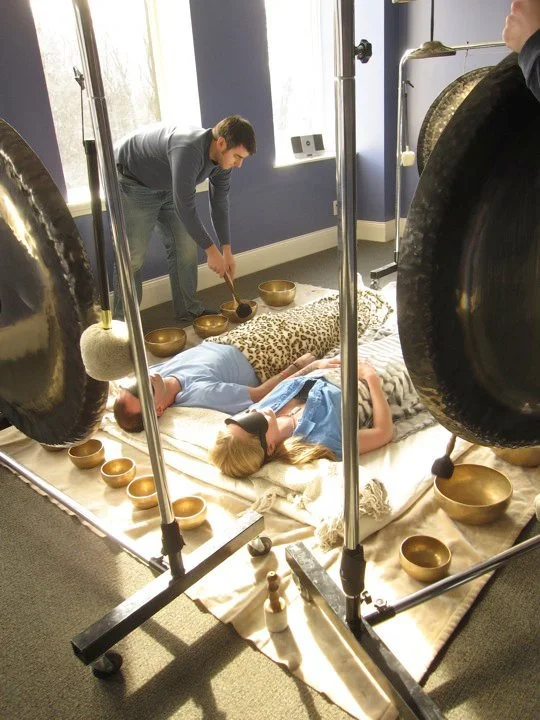 A couple lying on the floor with a blindfold, surrounded by singing bowls, as another person rings a bowl with a mallet in The Conduit Center's healing room with blue walls and large windows.