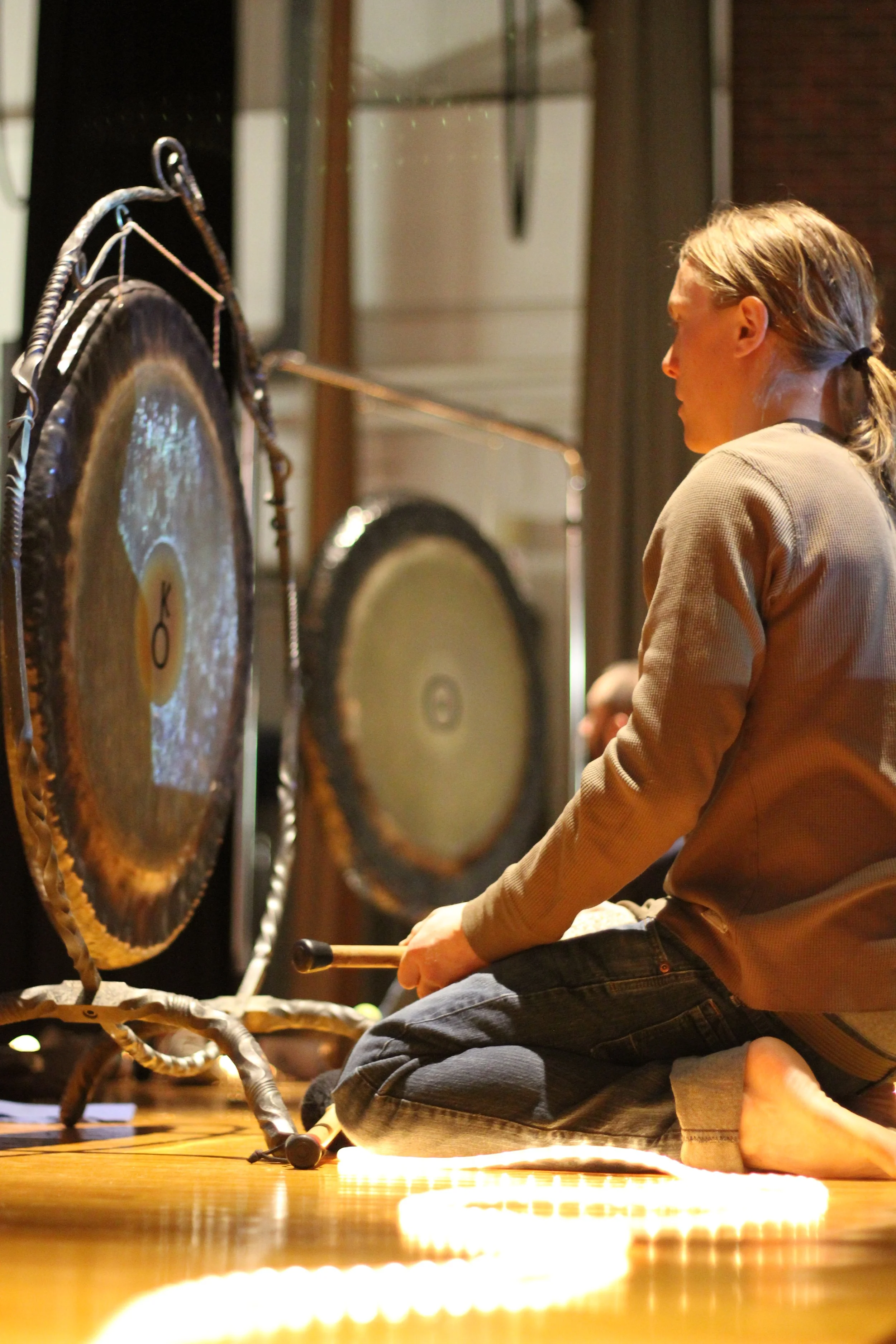 A woman kneeling on the floor playing two large gongs with mallets during a musical performance.