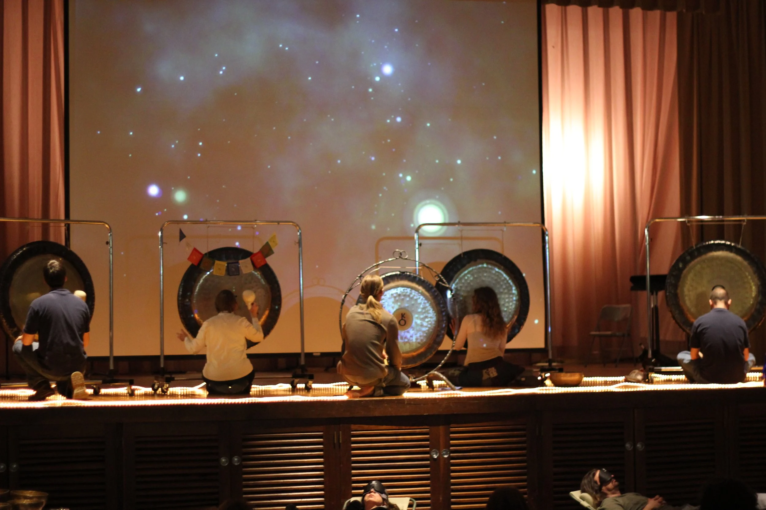 Five people kneeling on stage playing large gongs in front of a starry sky projection