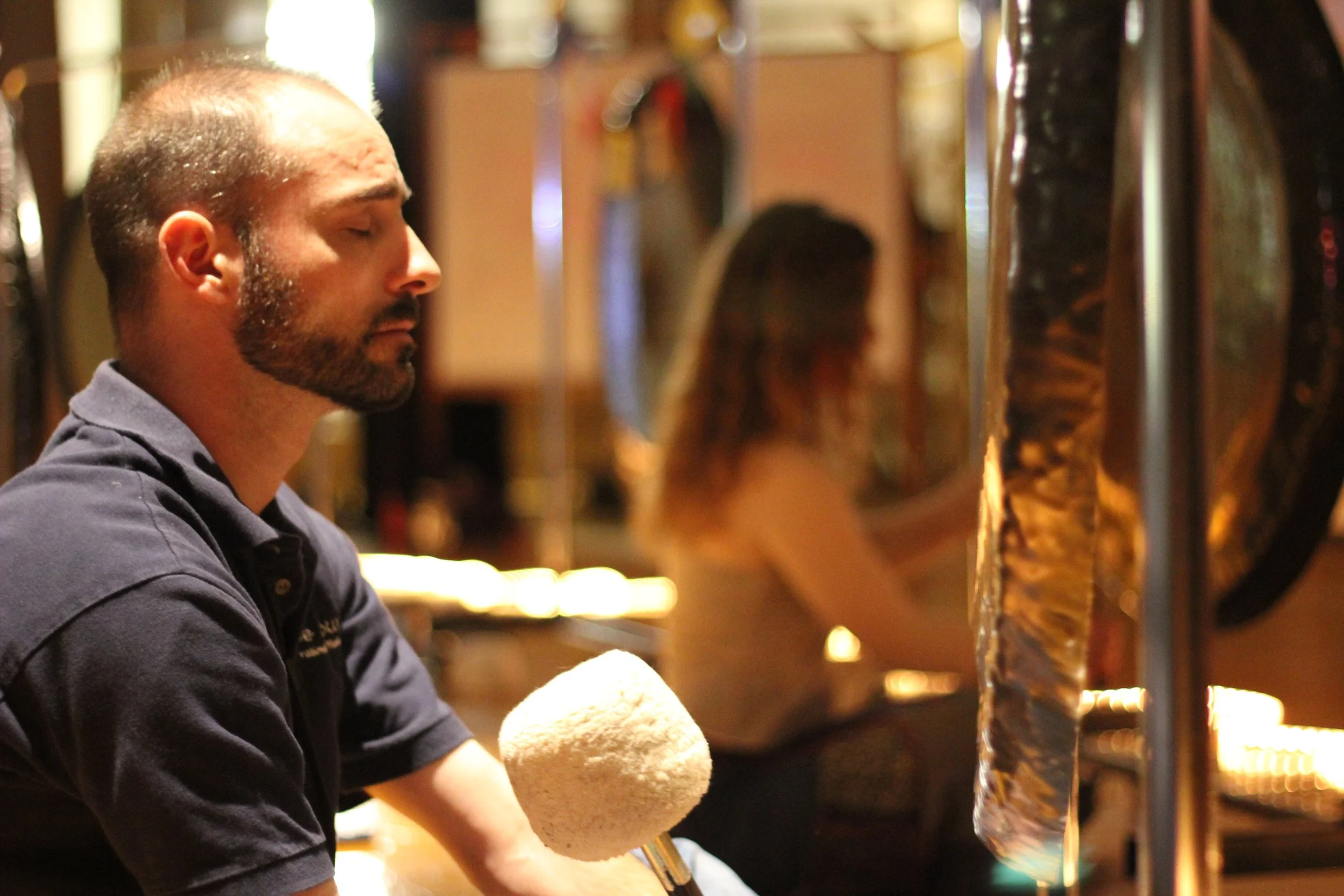A man with closed eyes and a beard playing a gong in a dimly lit room, with a woman in the background also near a gong.
