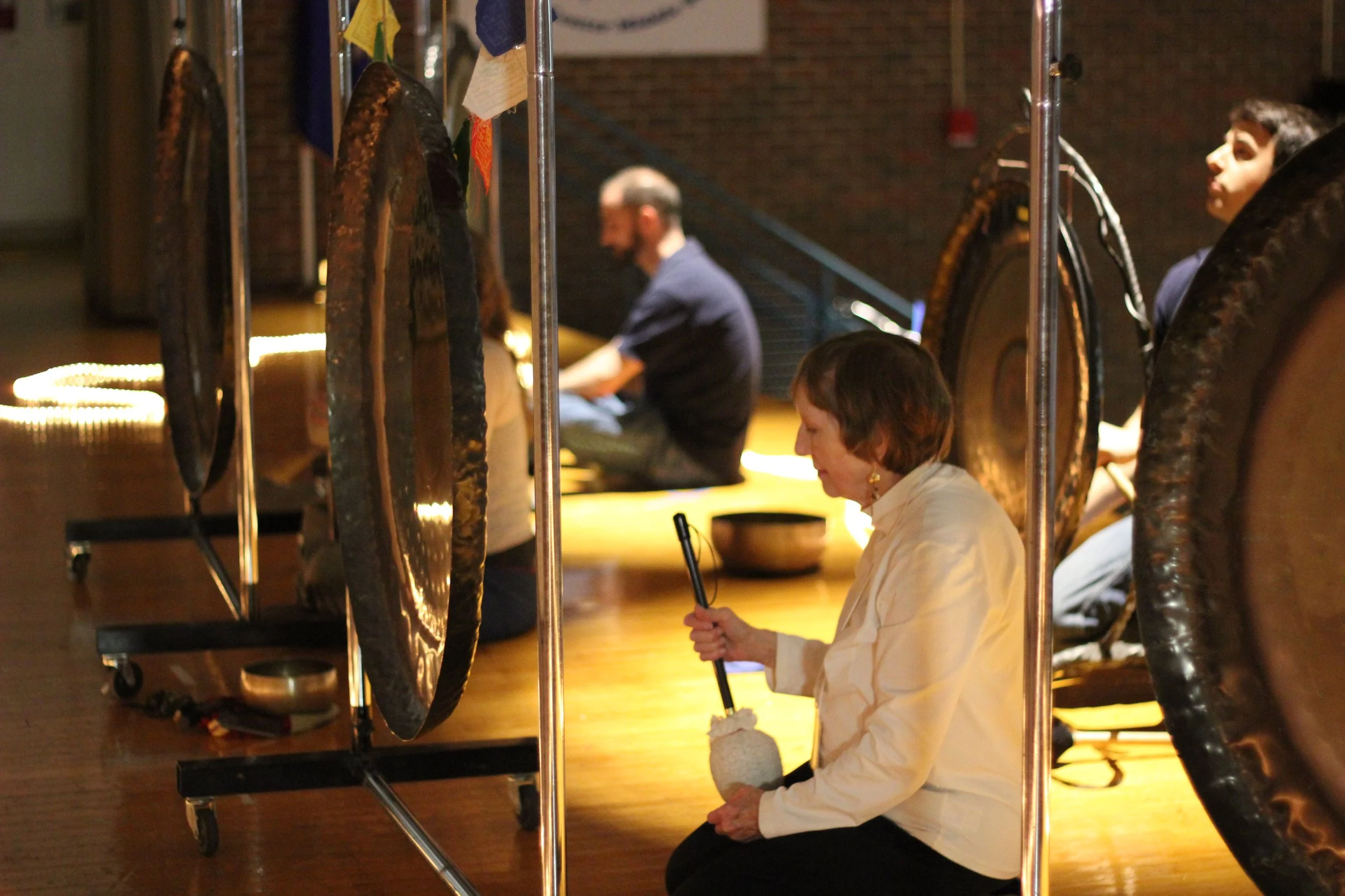 Three people sitting on the floor with large hanging gongs in front of them, possibly meditating or performing a sound healing session in a dimly lit room.