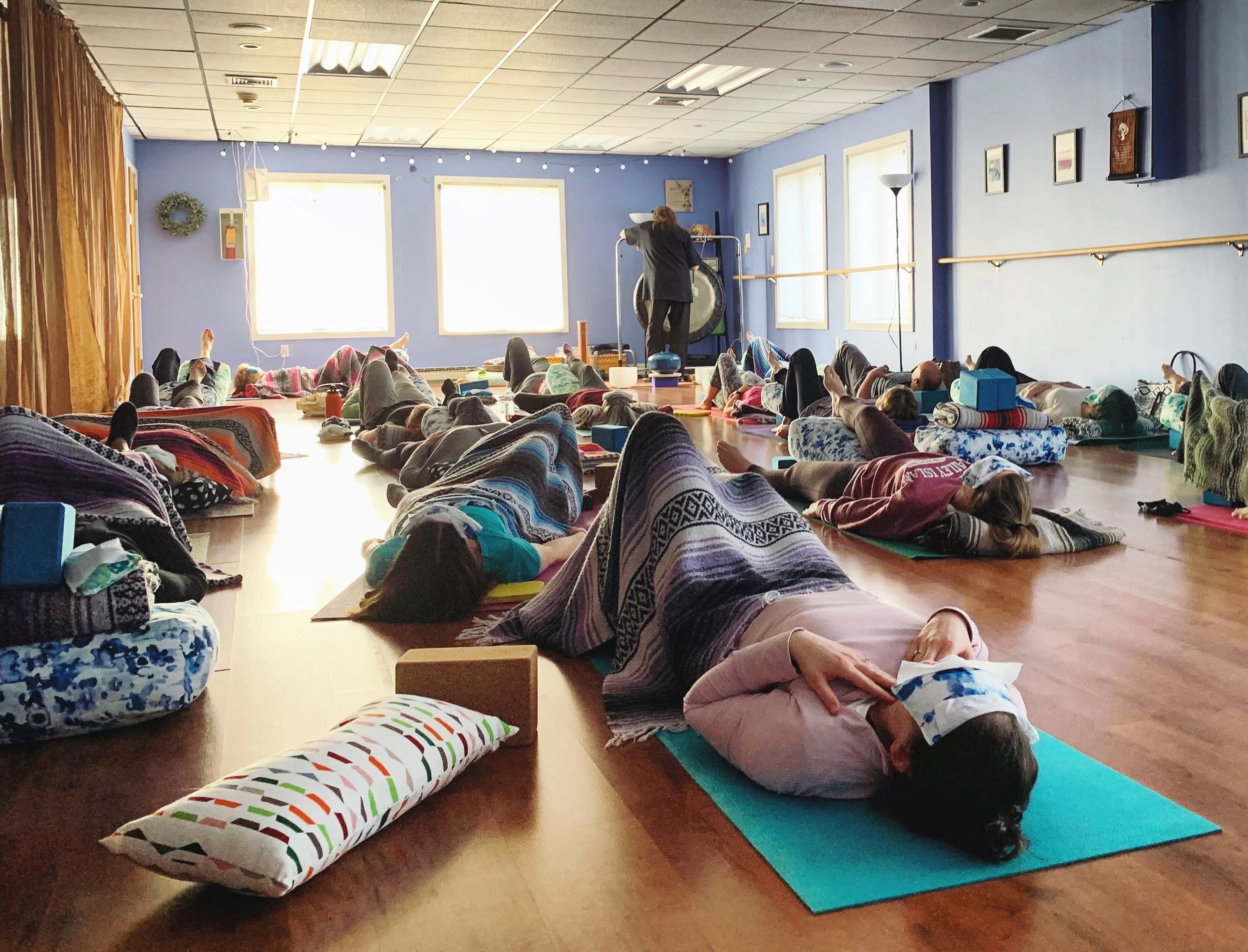 A group of people practicing yoga in a studio with large windows and blue walls, with a person playing a gong at the front.