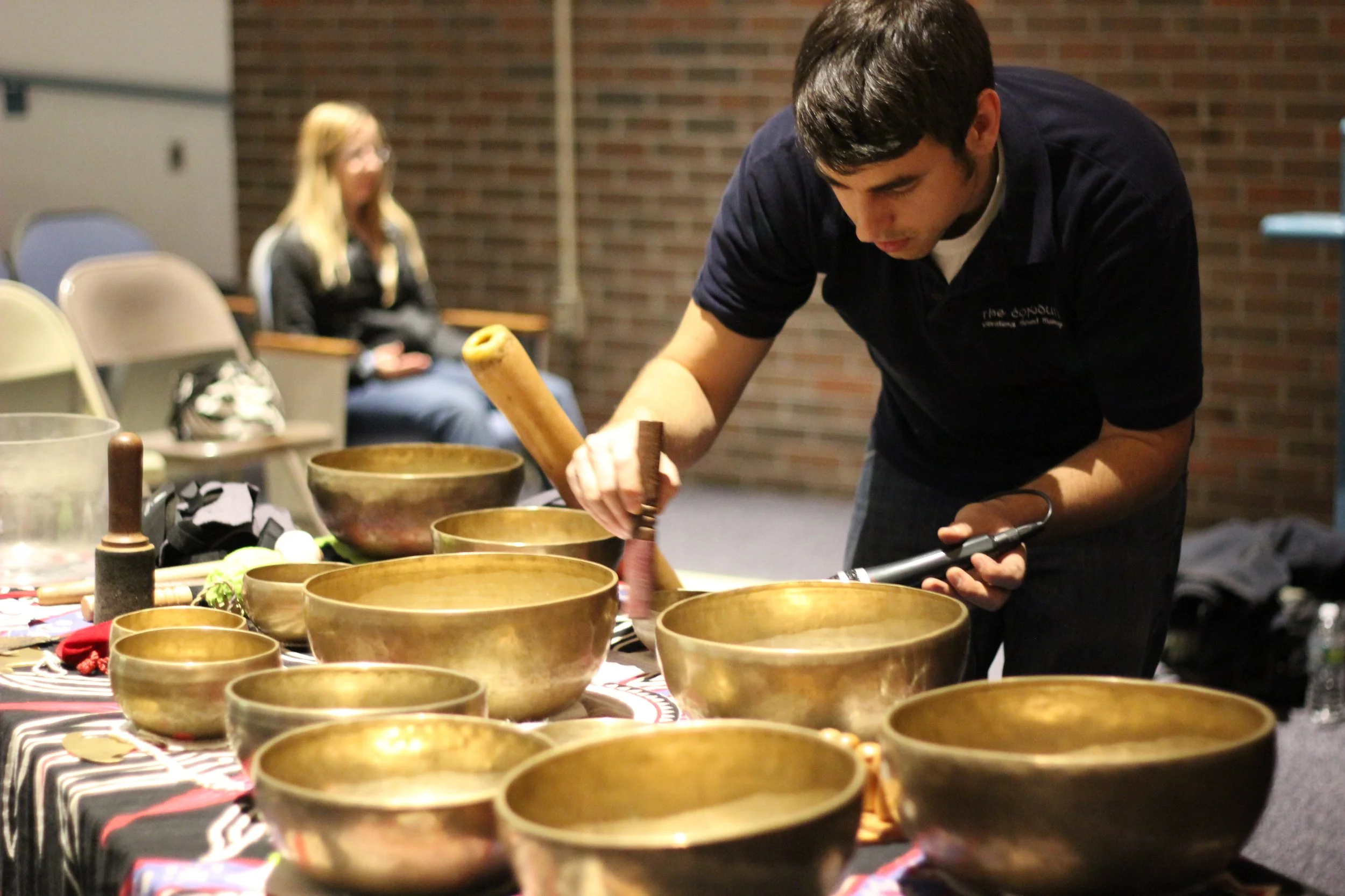 A man is playing singing bowls on a table, with a woman sitting in the background observing, in an indoor setting with a brick wall.