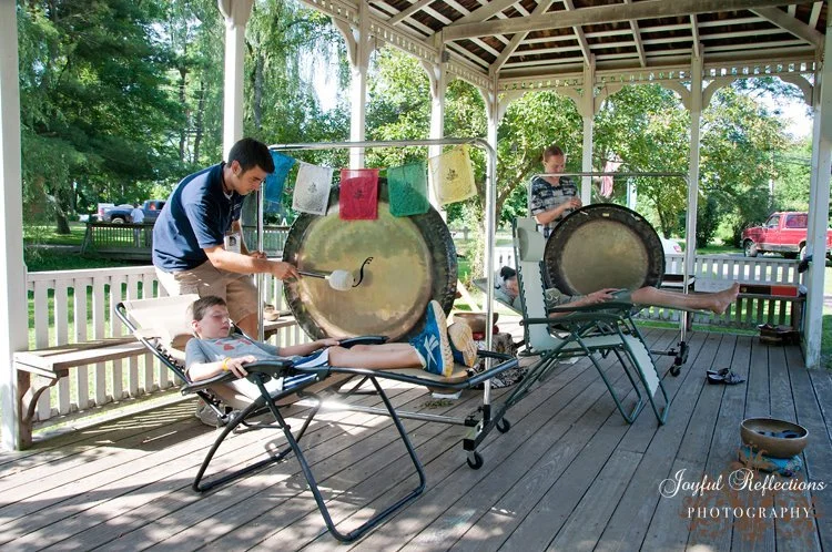 Two children receiving a gong healing demonstration lounging in zero-gravity chairs with two Conduit musicians playing large gongs under a gazebo on a porch, surrounded by trees.