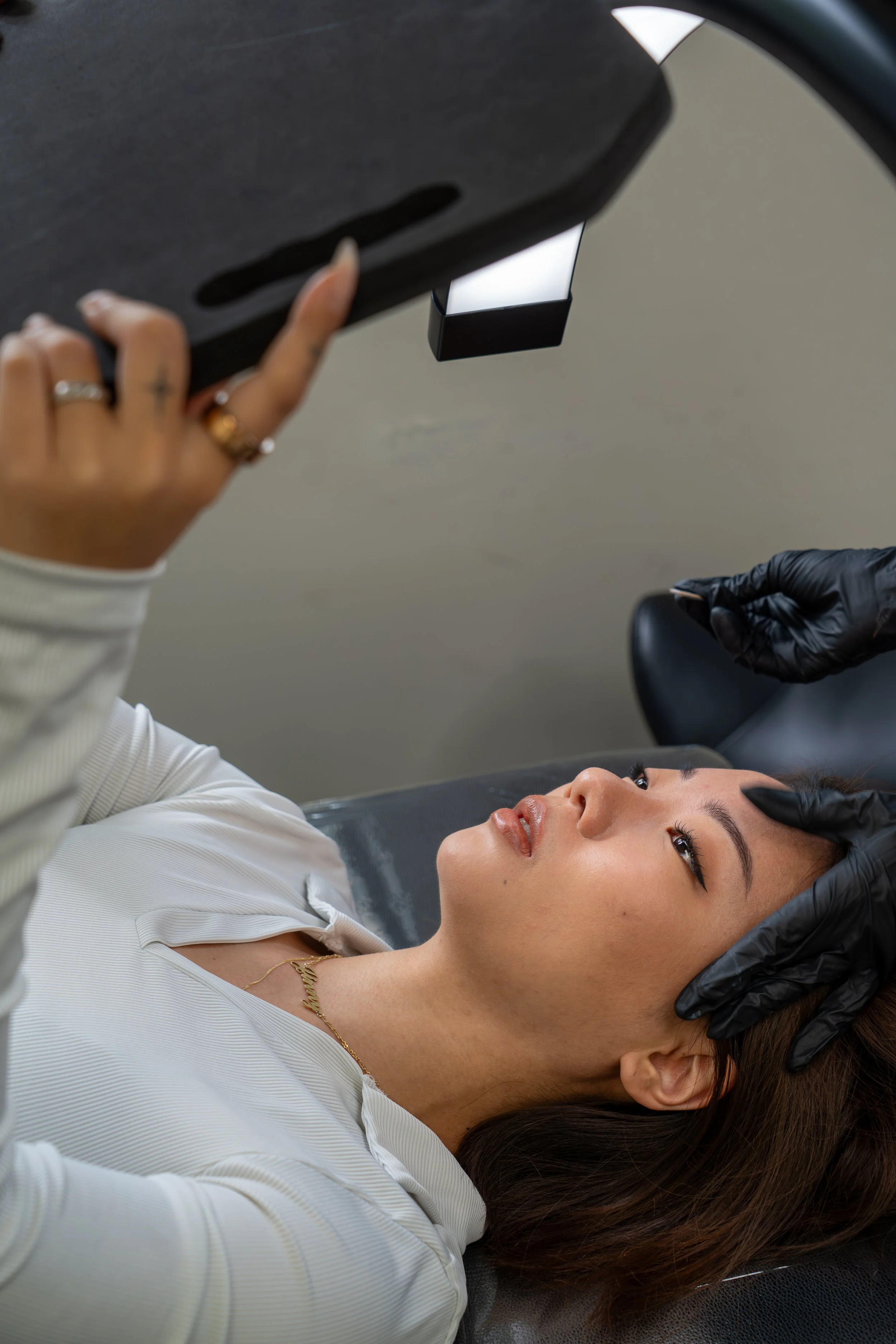 A woman lying on a treatment chair, receiving a skincare treatment. A person wearing black gloves is holding her forehead while another person holds a device near her face.