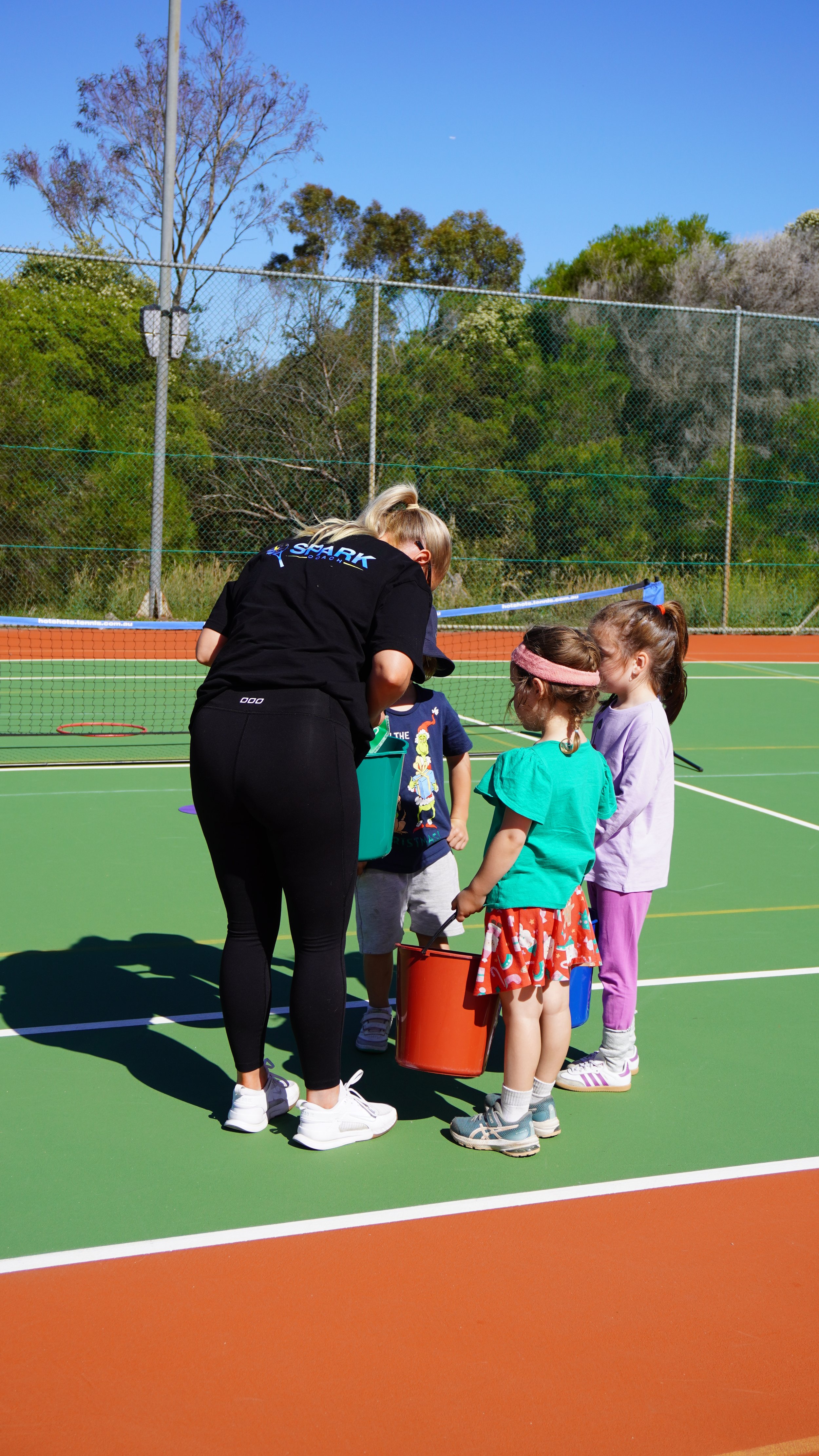 A woman coaching four young children on a tennis court during daytime with trees and a chain-link fence in the background.