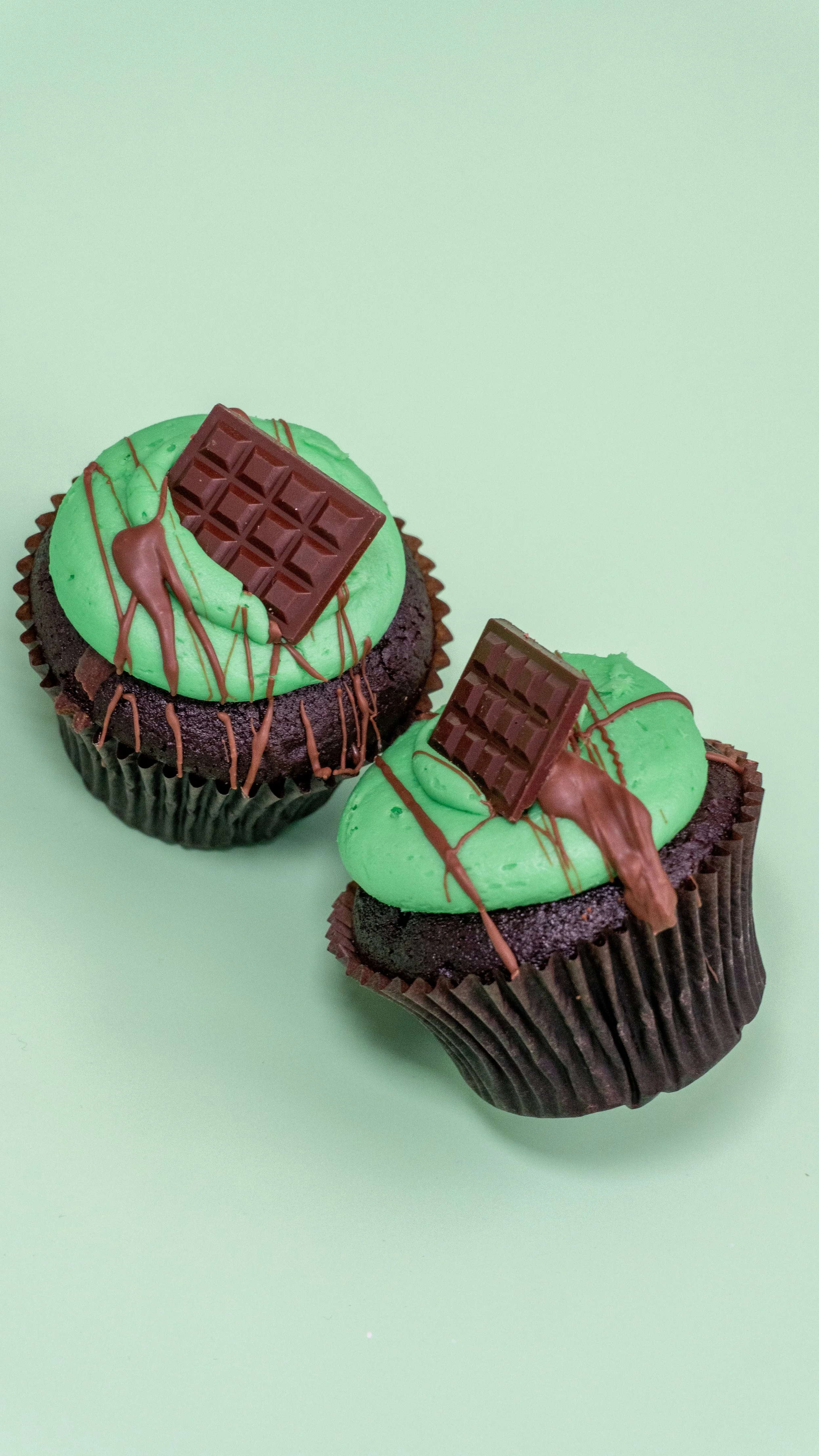 Two chocolate cupcakes with green frosting, topped with chocolate squares and drizzled with chocolate sauce, on a mint green background.