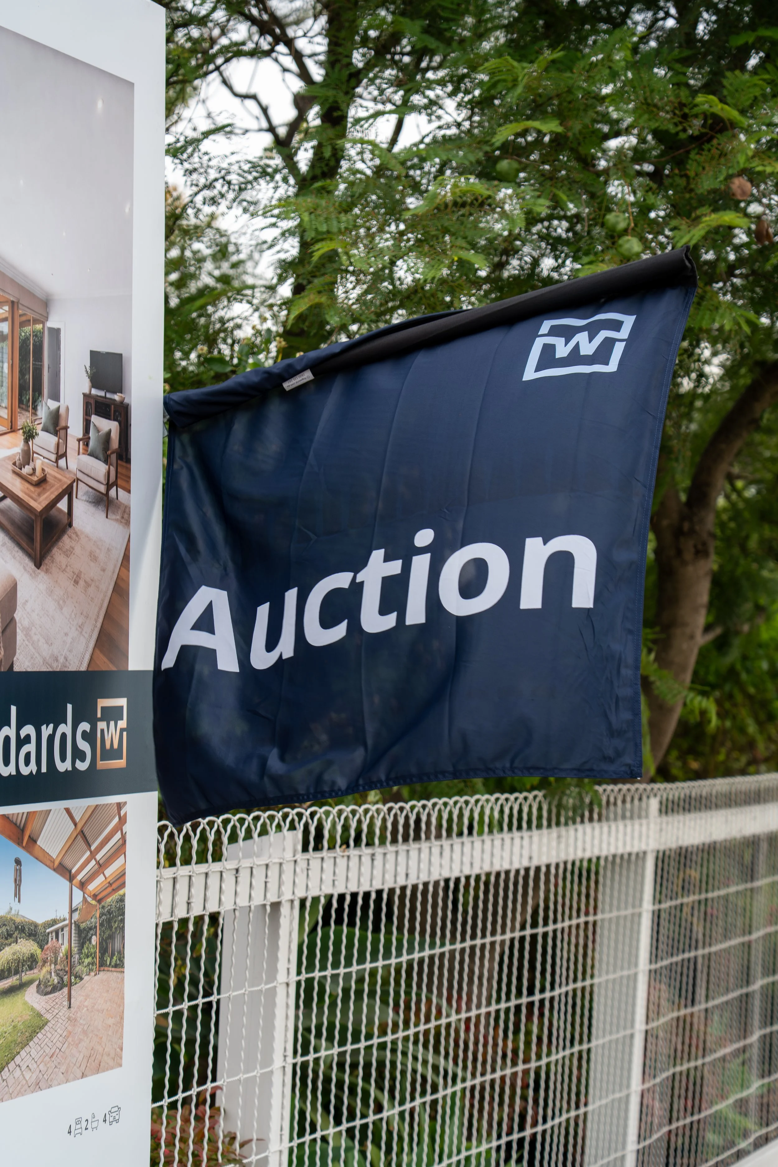 A black flag with white text that reads "Auction" is attached to a white chain-link fence. The fence is in front of a background of green trees and a residential neighborhood. An advertisement poster for a real estate auction is partly visible on the left side of the image.