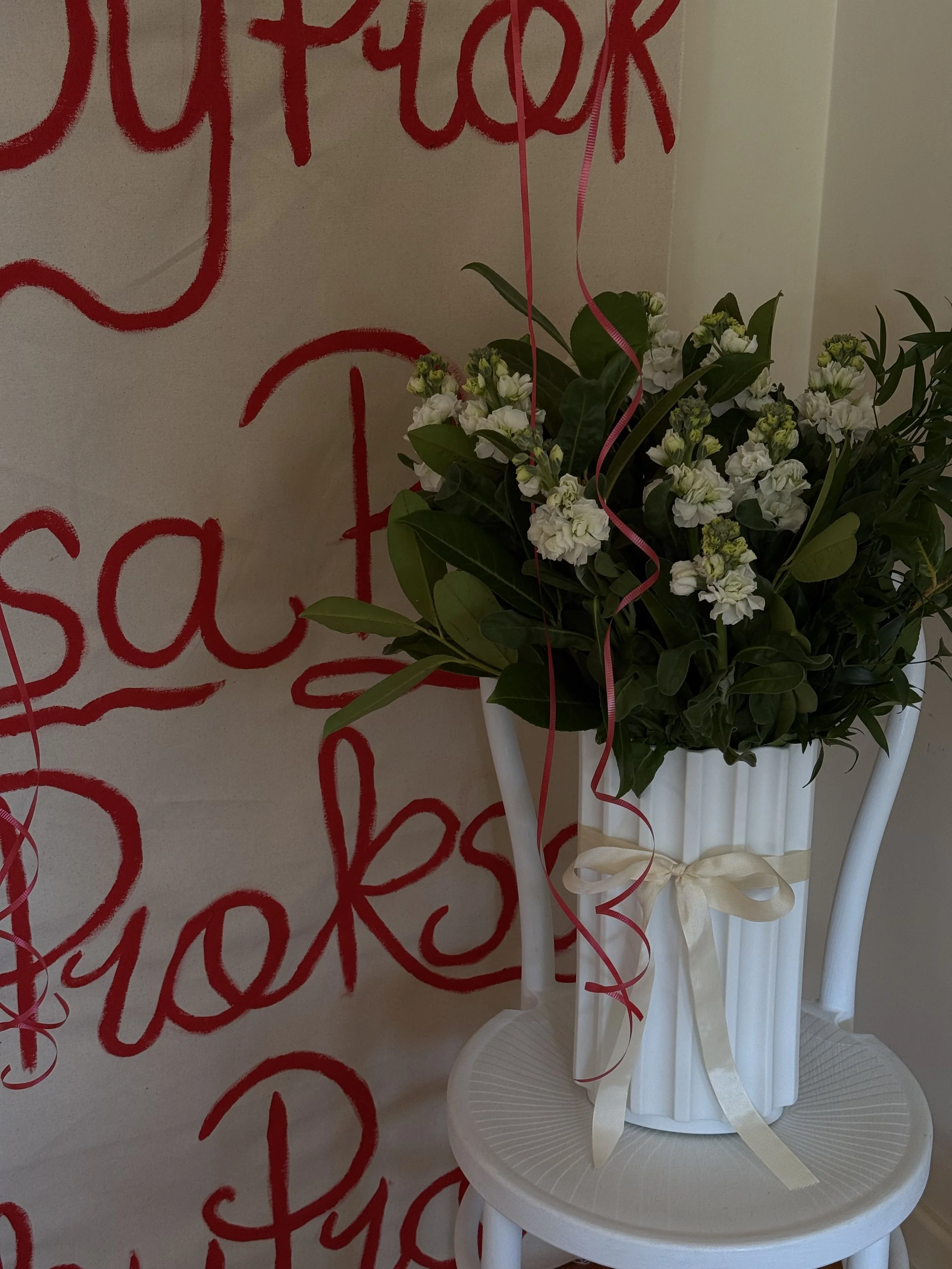 A white vase with a cream ribbon tied in a bow, filled with white flowers and green leaves, placed on a white chair against a wall with red handwritten-style text.