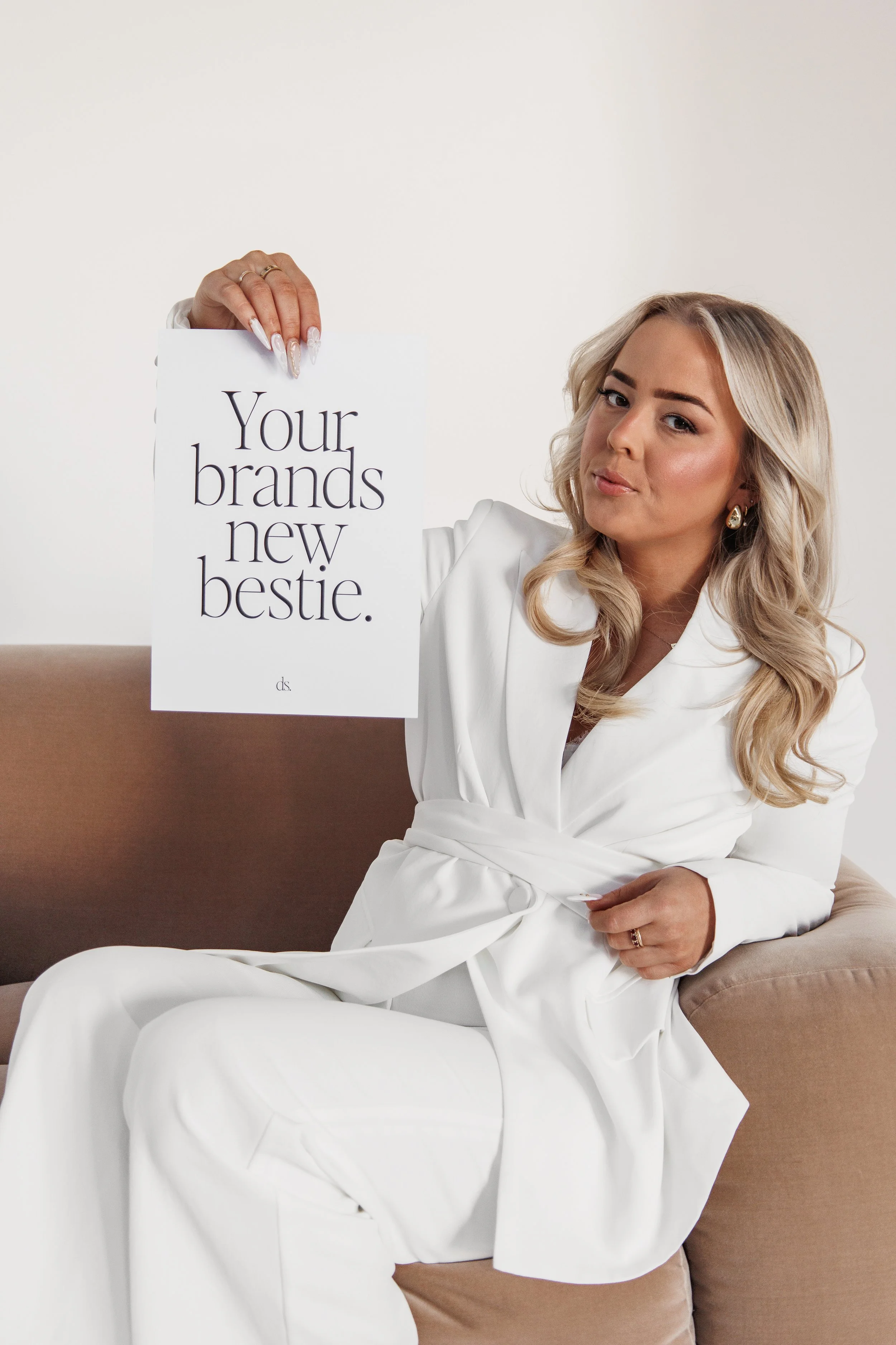 A woman in a white outfit sitting on a beige couch, holding a white sign that reads "Your brands new bestie."