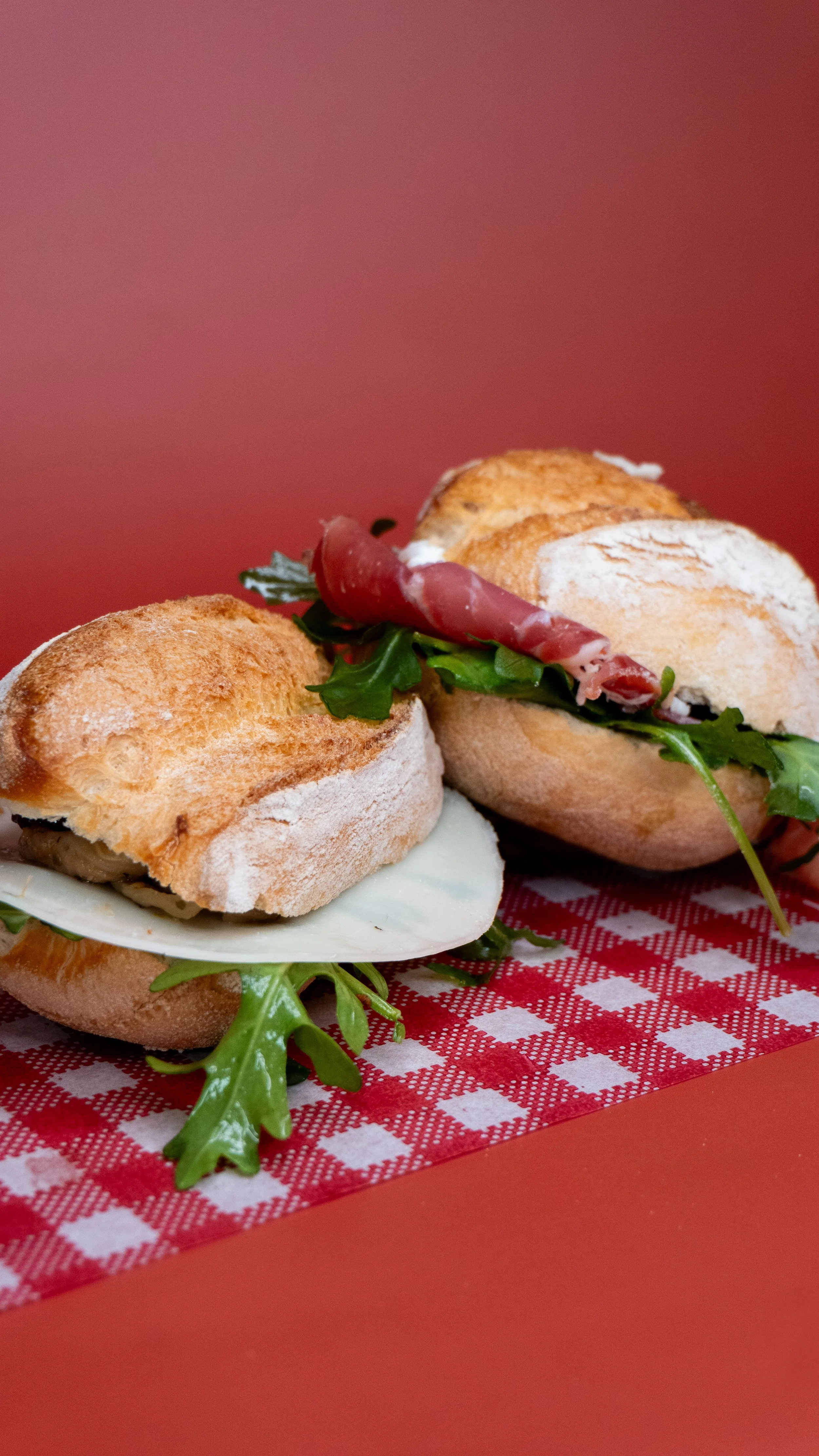 Two sandwiches with prosciutto, arugula, cheese, and bread rolls, placed on a red and white checkered paper on a red table