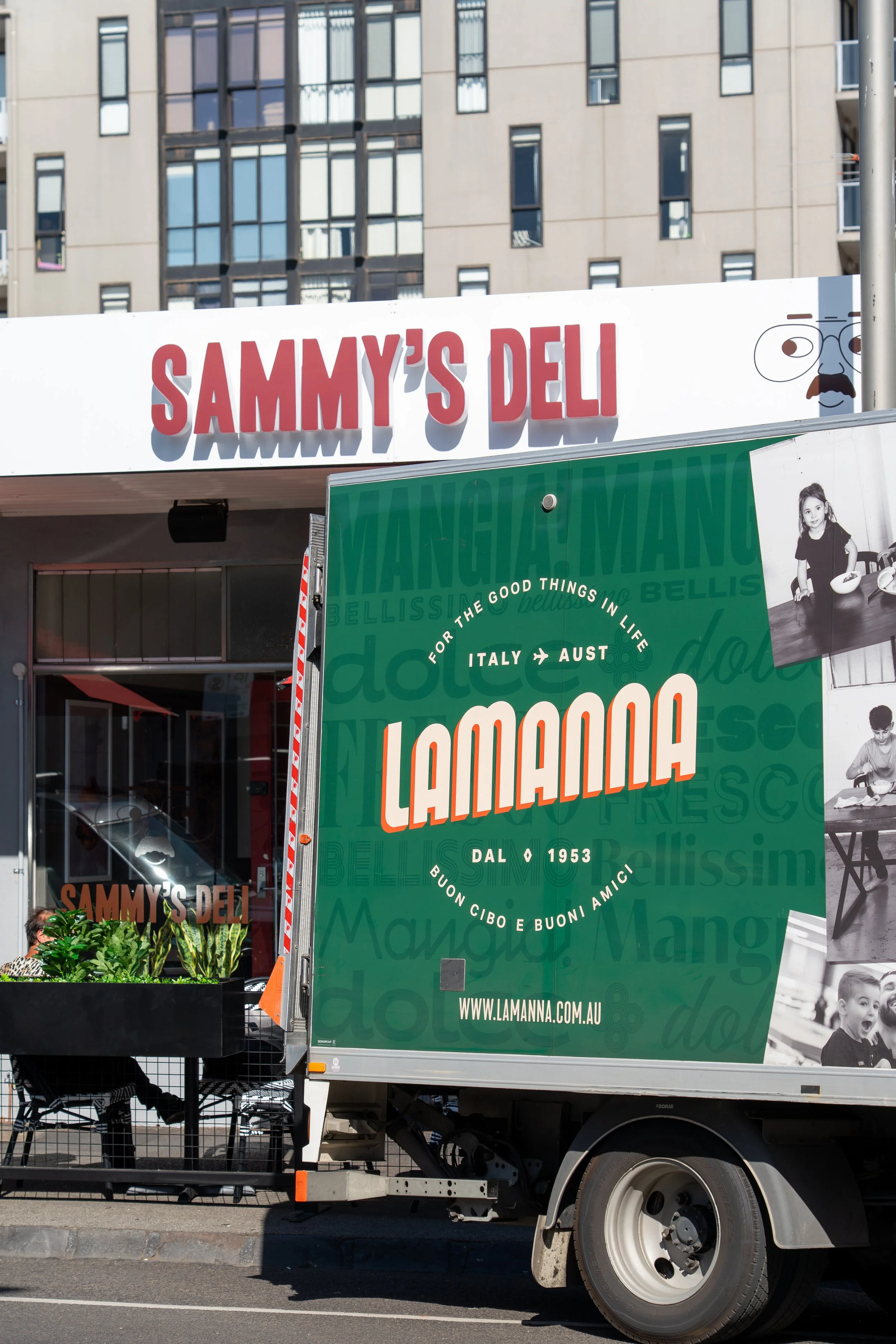 A food truck with signage for Sammy's Deli and Lamanna, featuring green and white color themes, located on a city street with residential buildings in the background.