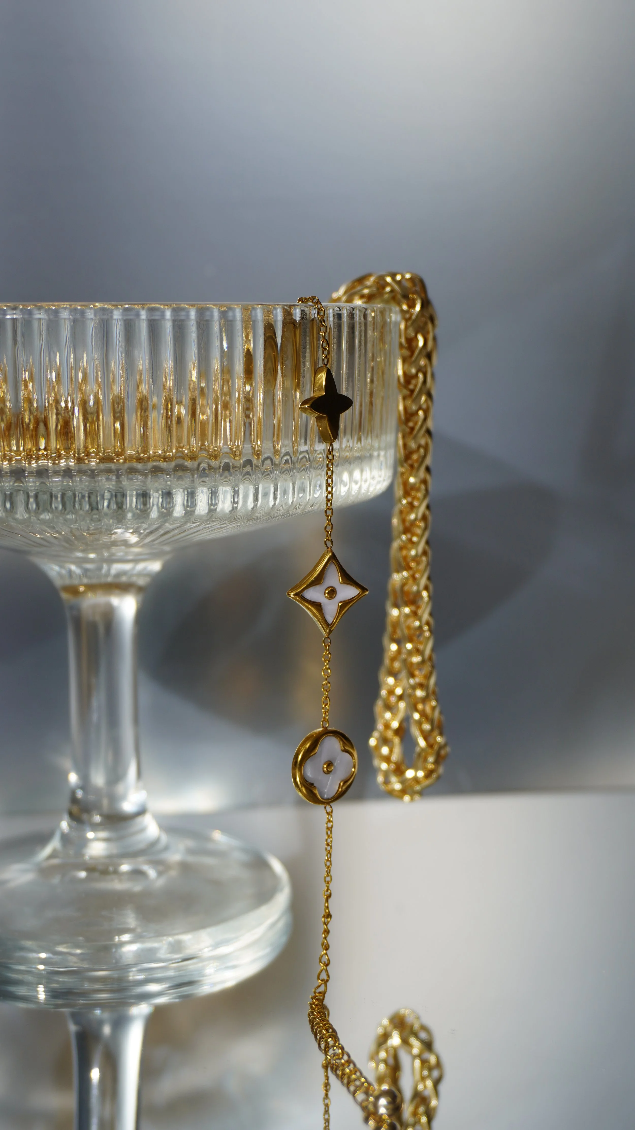 Gold jewelry hanging from the rim of a glass bowl with a chain and gold bracelet draped over it, against a light background.