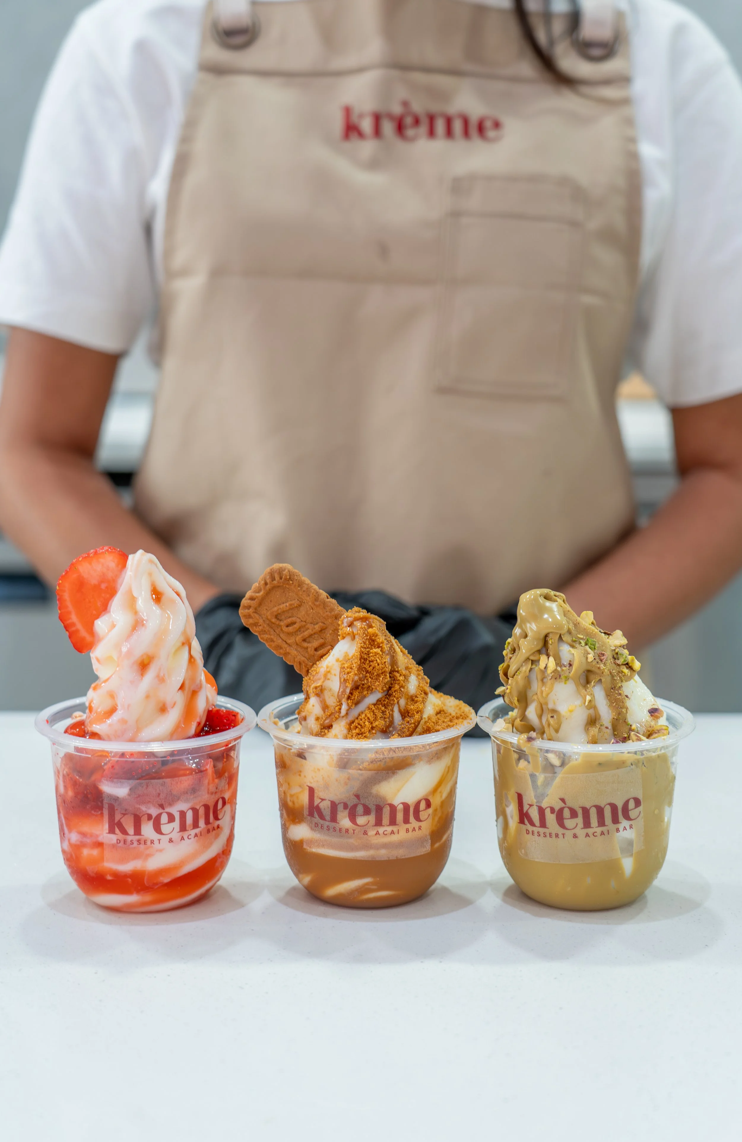 Three cups of ice cream with toppings from Kreme, a dessert and acai bar, are placed on a white surface. The flavors appear to be strawberry shortcake, caramel, and pistachio, each with various toppings. A person wearing a beige apron with the 'kreme' logo is standing behind the cups.