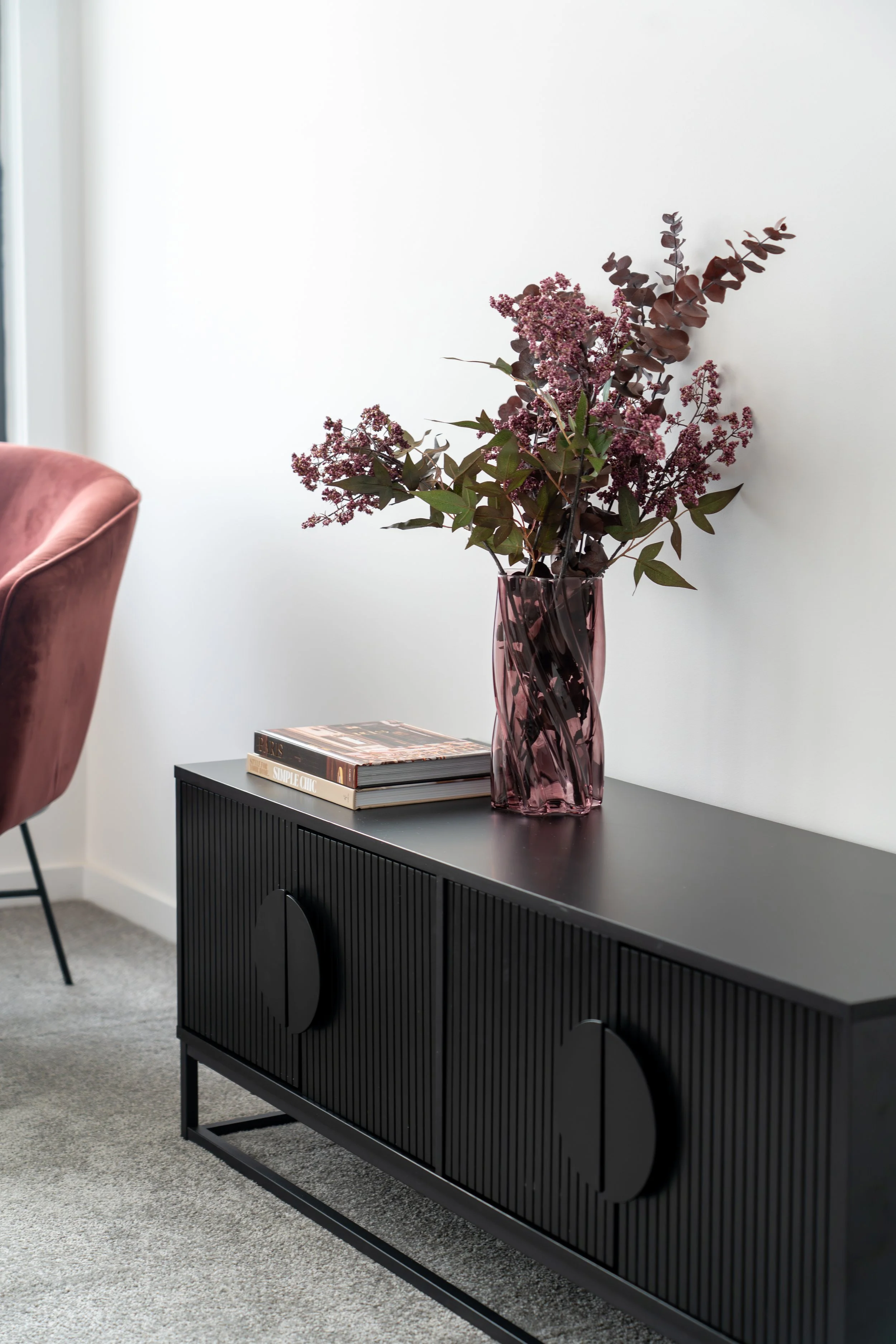 A black sideboard with a textured front and round cutouts in the doors, set against a white wall. On top of the sideboard, there is a pink glass vase containing a bouquet of purple and burgundy flowers. Next to the vase, there are three stacked books. To the left, part of a pink velvet armchair is visible, and the floor is covered in a light gray carpet.