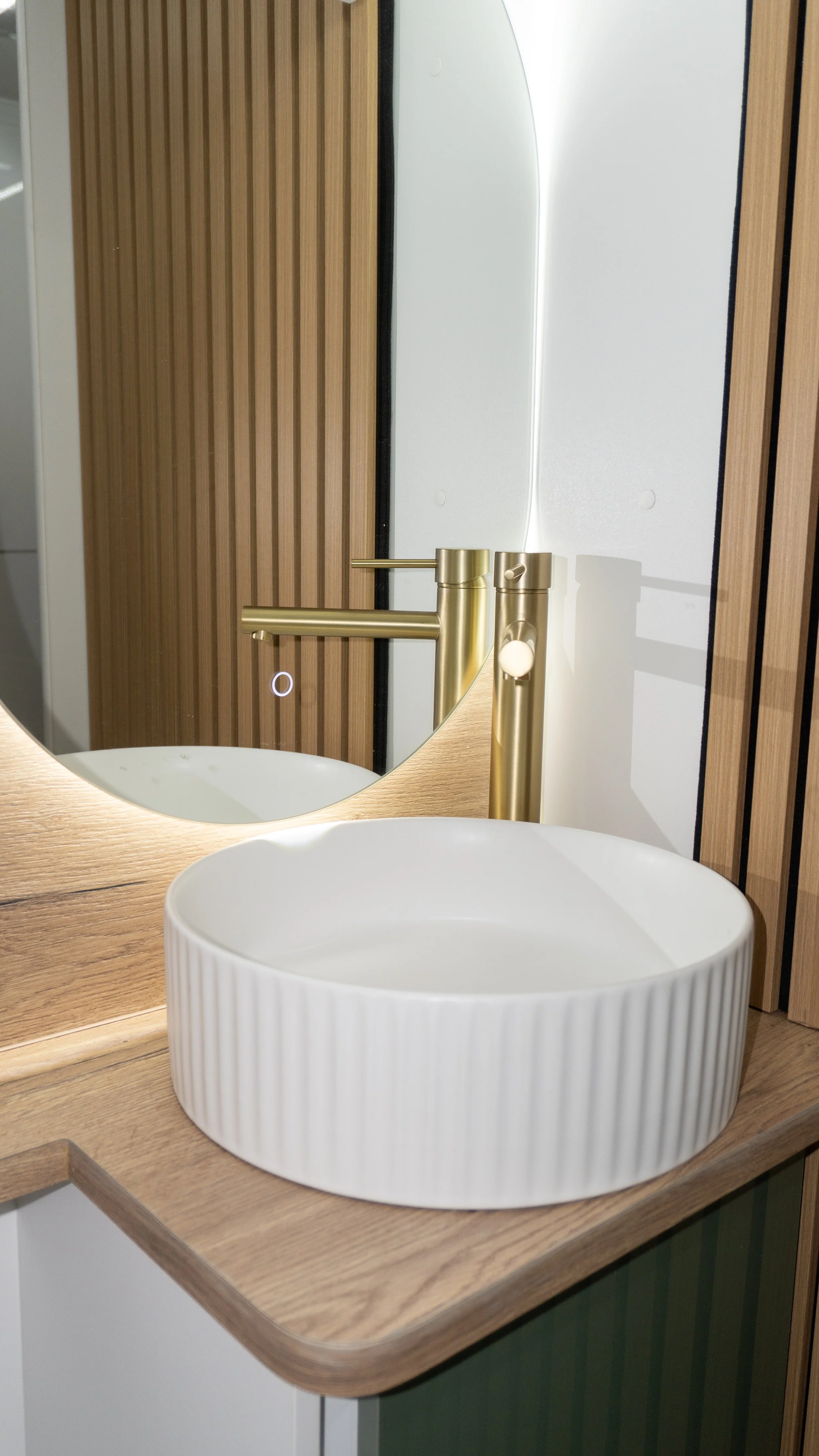 A bathroom sink with a round, white, ridged ceramic basin on a wooden countertop, with a large mirror reflecting a gold faucet and part of a wooden wall.