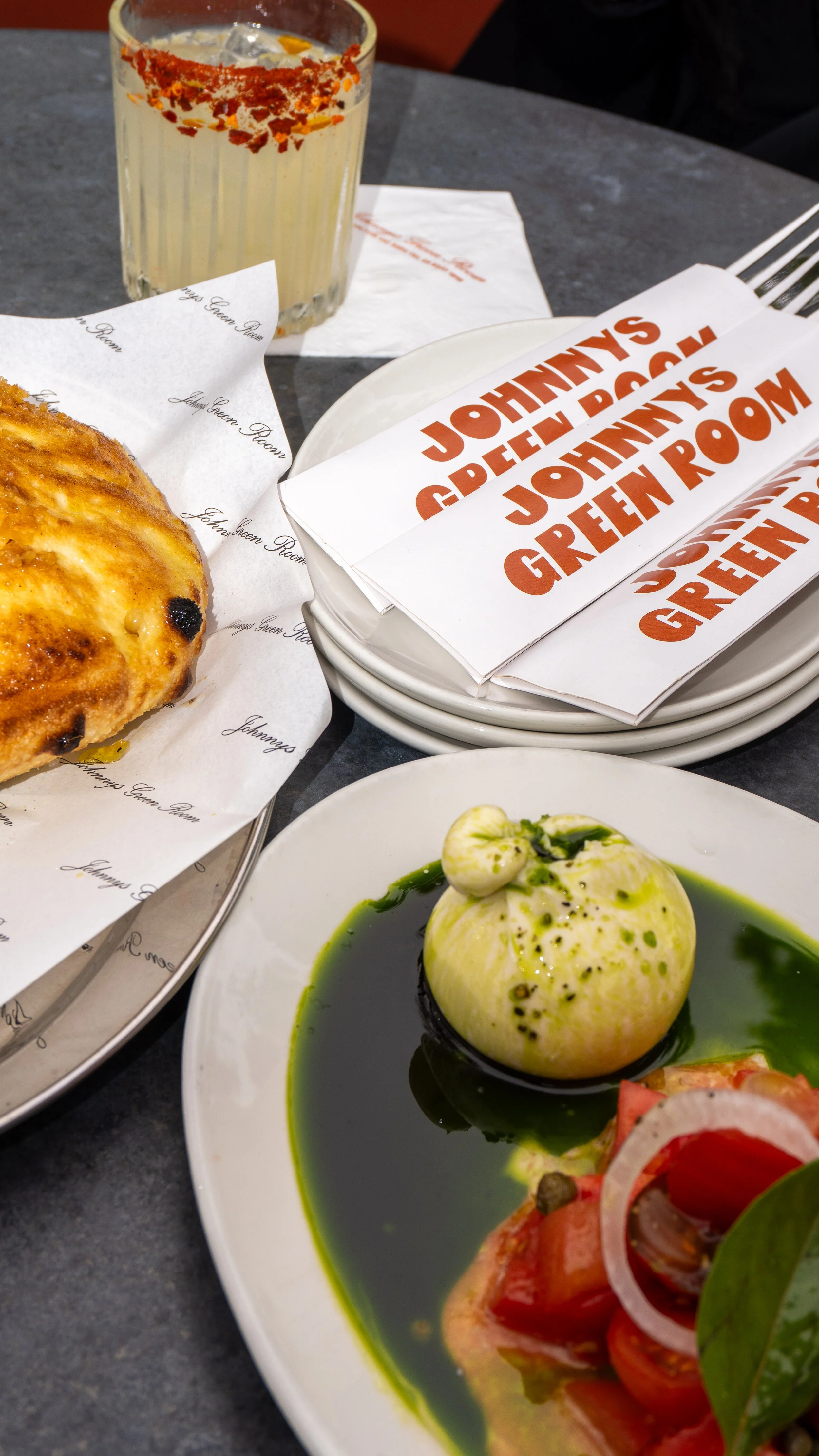 A plate with a round cheese ball topped with black pepper, served in a green oil-based sauce. In the background, there is a glass of a light-colored beverage with red seasoning around the rim, and a partially visible pastry and a paper with text reading 'JOHNNY GREEN ROOM'.