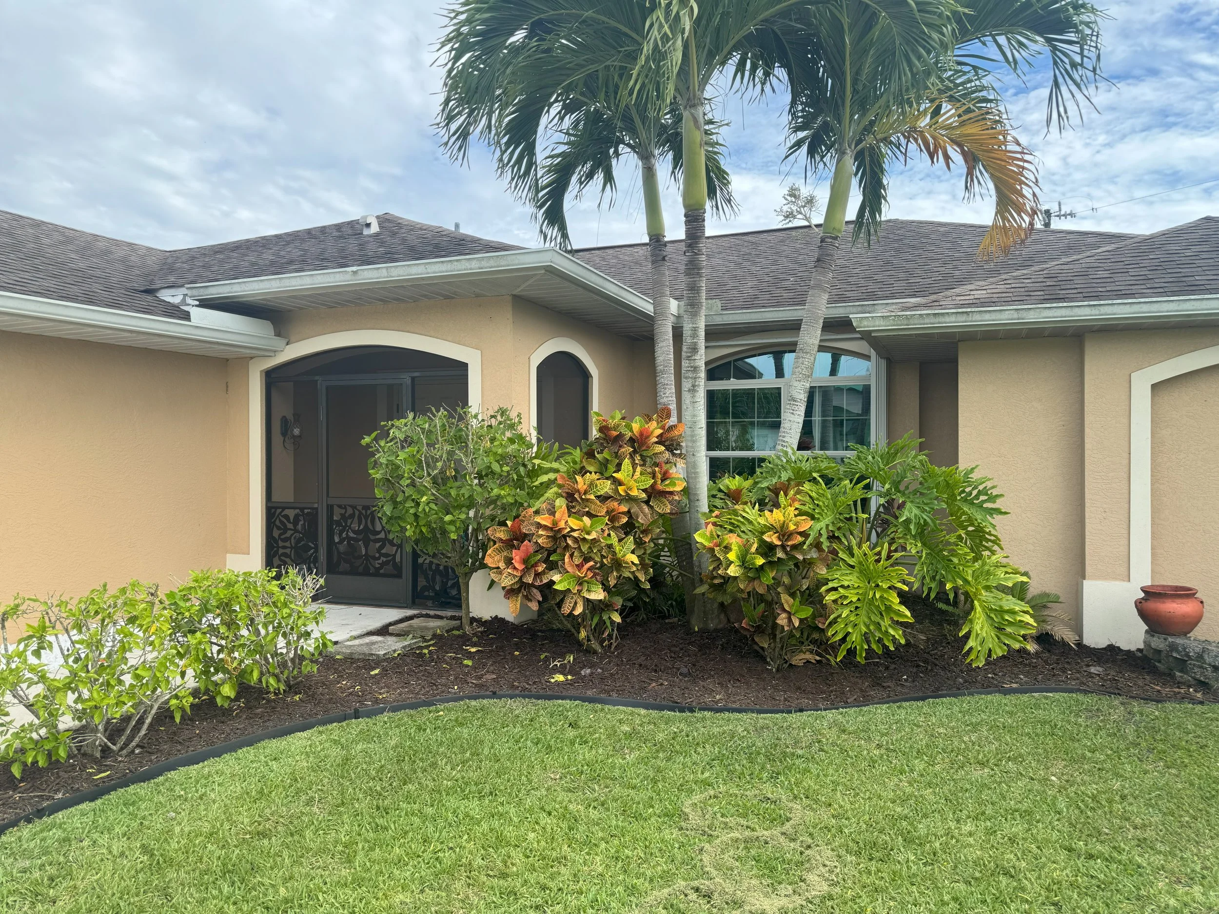 A house with beige exterior walls, dark roof, and multiple windows. In front, there's a well-maintained lawn, some bushes, and two tall palm trees. A potted plant is placed near the right corner.