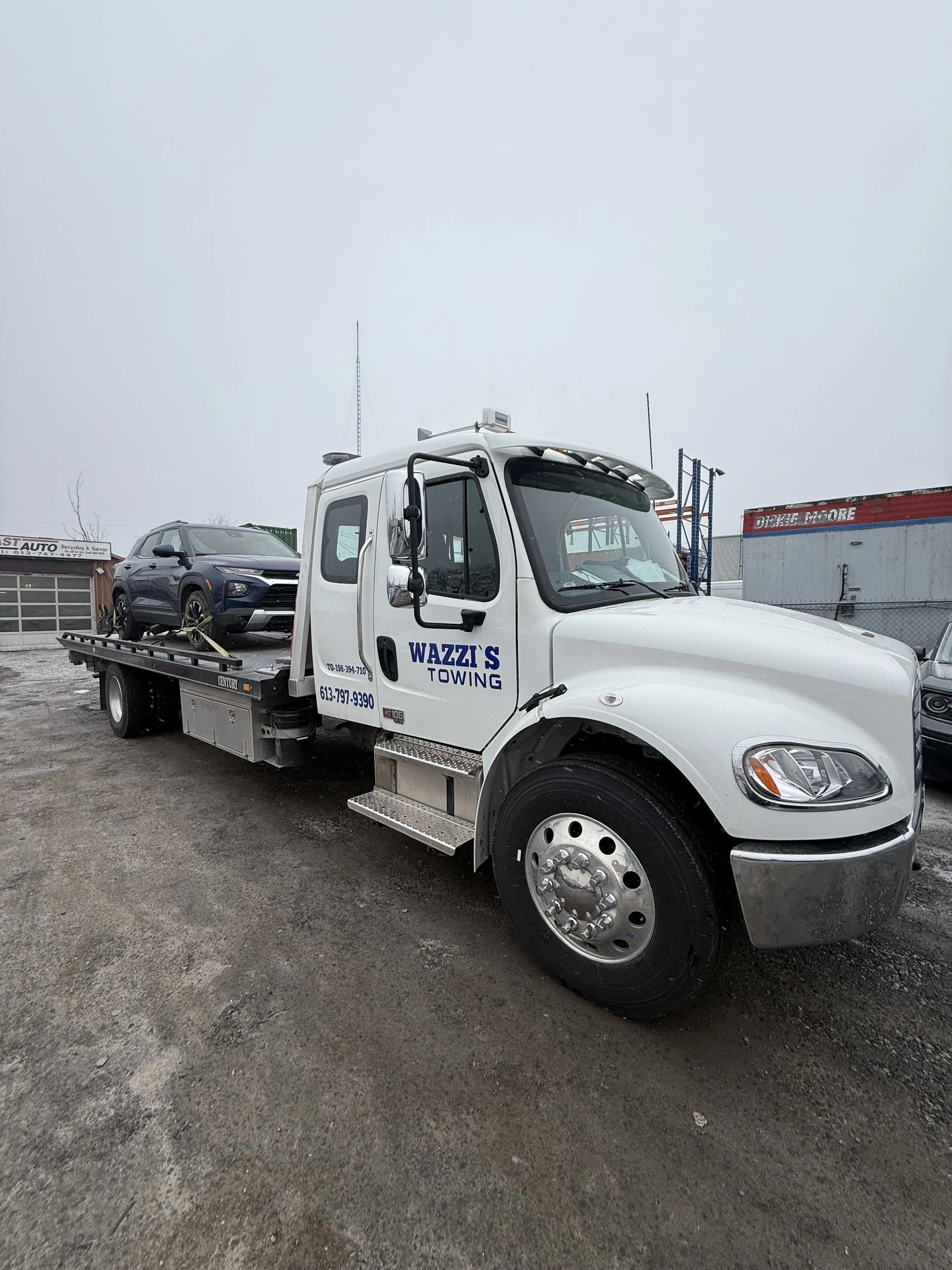 A white tow truck with the words "Wazzi's Towing" on the door, lifting a black SUV in a lot with other vehicles and industrial buildings in the background.