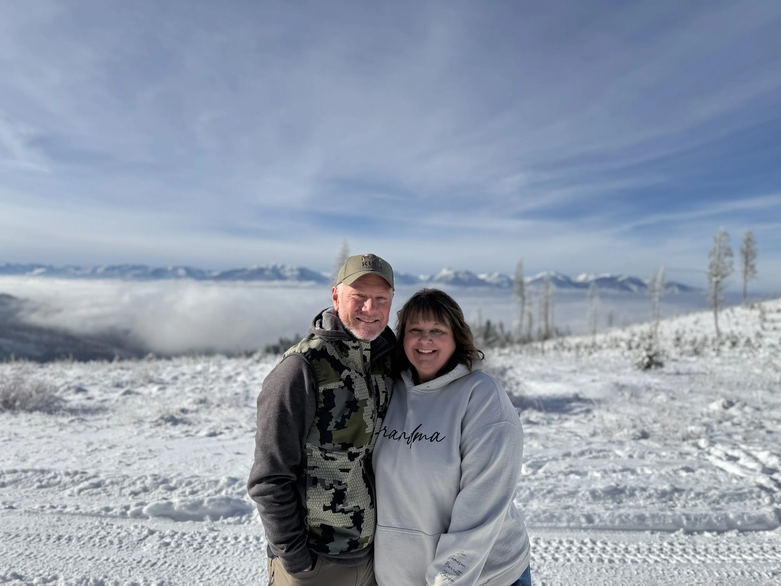 A smiling couple standing on snowy ground with a mountain range and cloudy sky in the background.