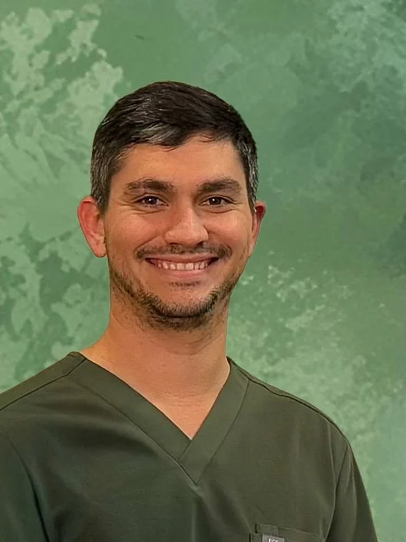 Man with curly dark hair, glasses, and a beard, wearing a dark green sweater, sitting indoors with a plant in the background.