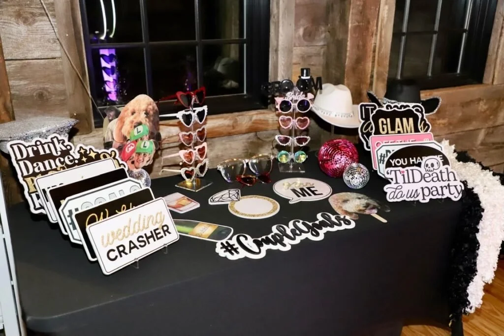 Photo of a party photo booth table decorated with signs, glasses, and props, featuring a wooden wall background.