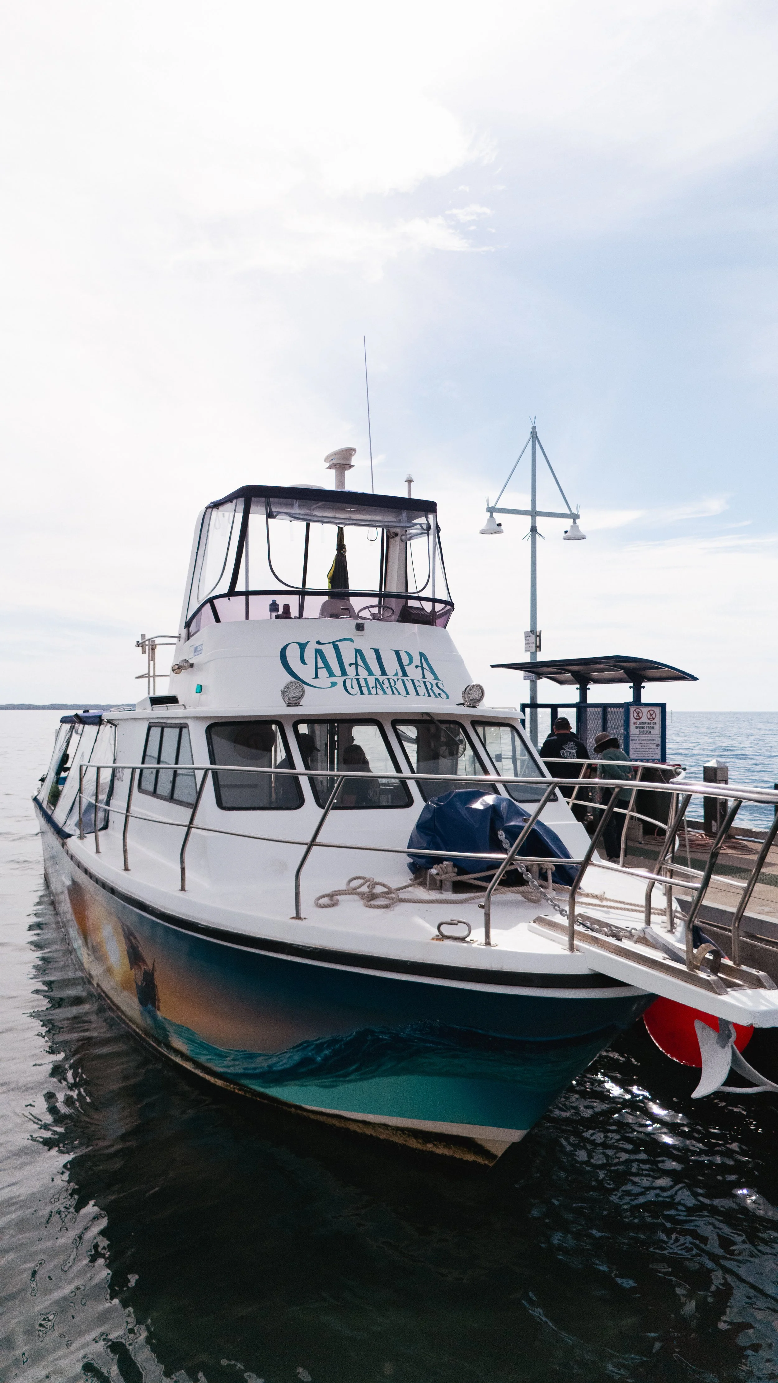 A white boat docked at a pier with the words "Catalpa Charters" on the front, and two people standing on the pier near the boat.