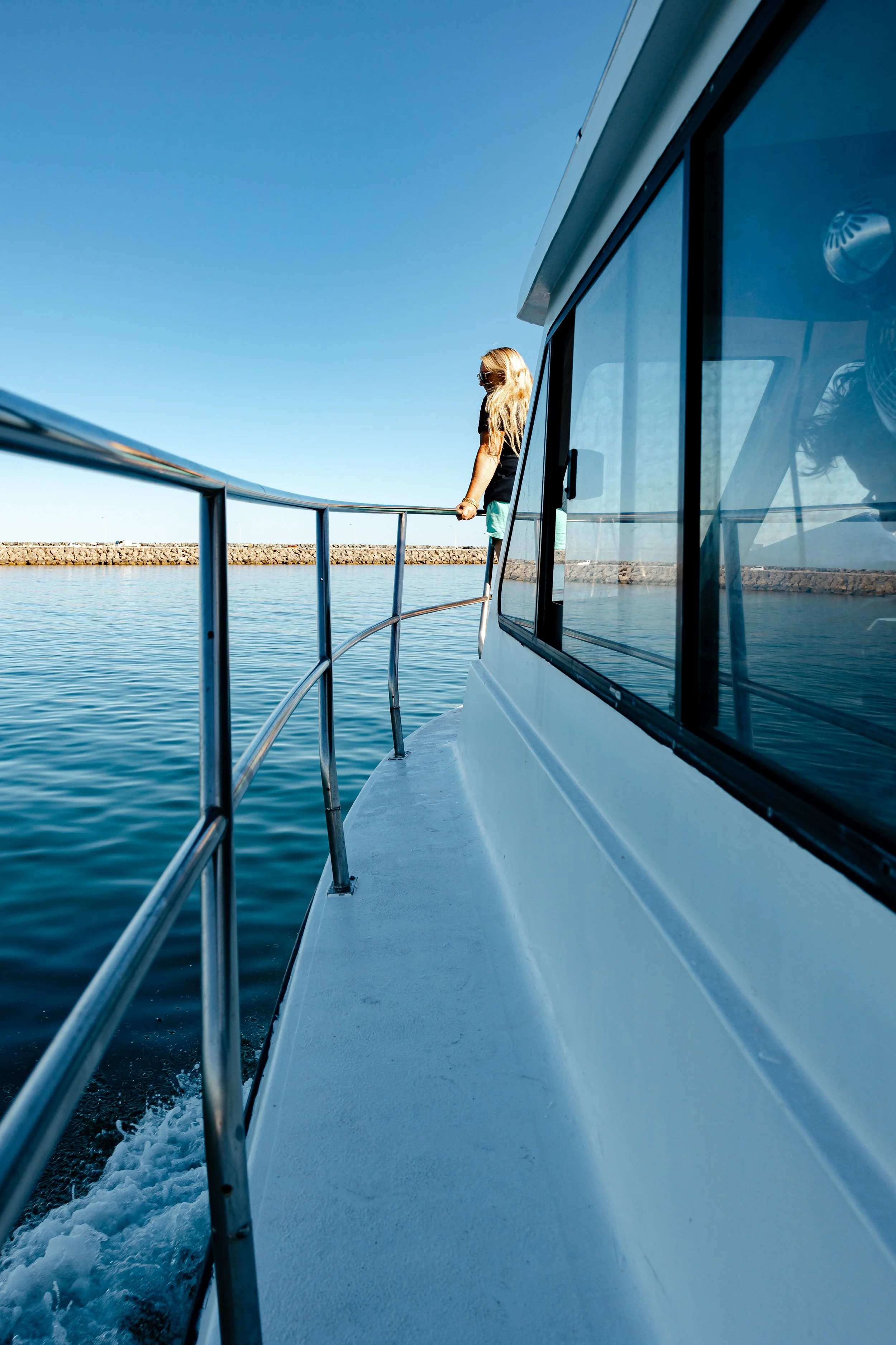 A woman with blonde hair, wearing sunglasses, black shirt, and turquoise shorts, standing on the deck of a boat, gazing out at the water with a clear blue sky in the background.