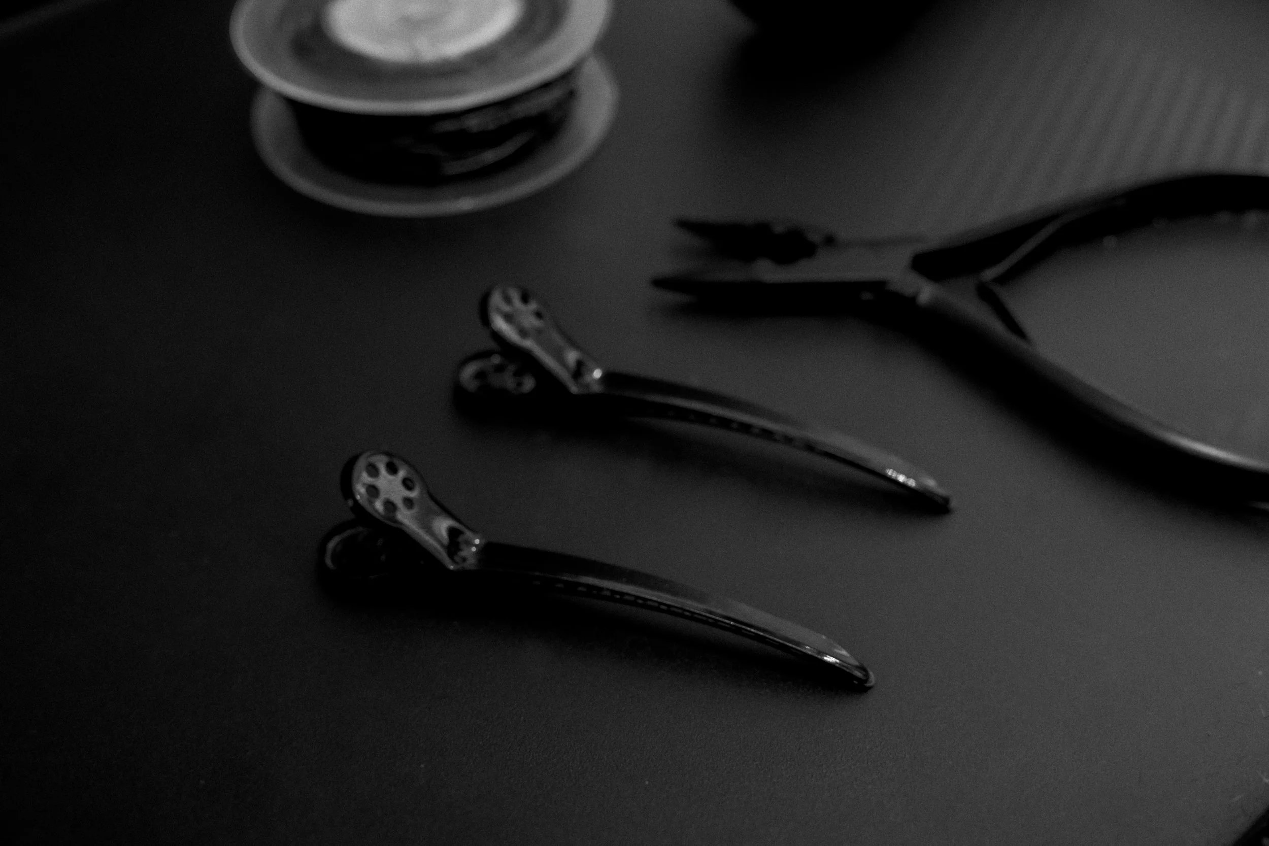 A pair of metal hair clips on a dark surface with a pair of scissors and small container of hairpins in the background.
