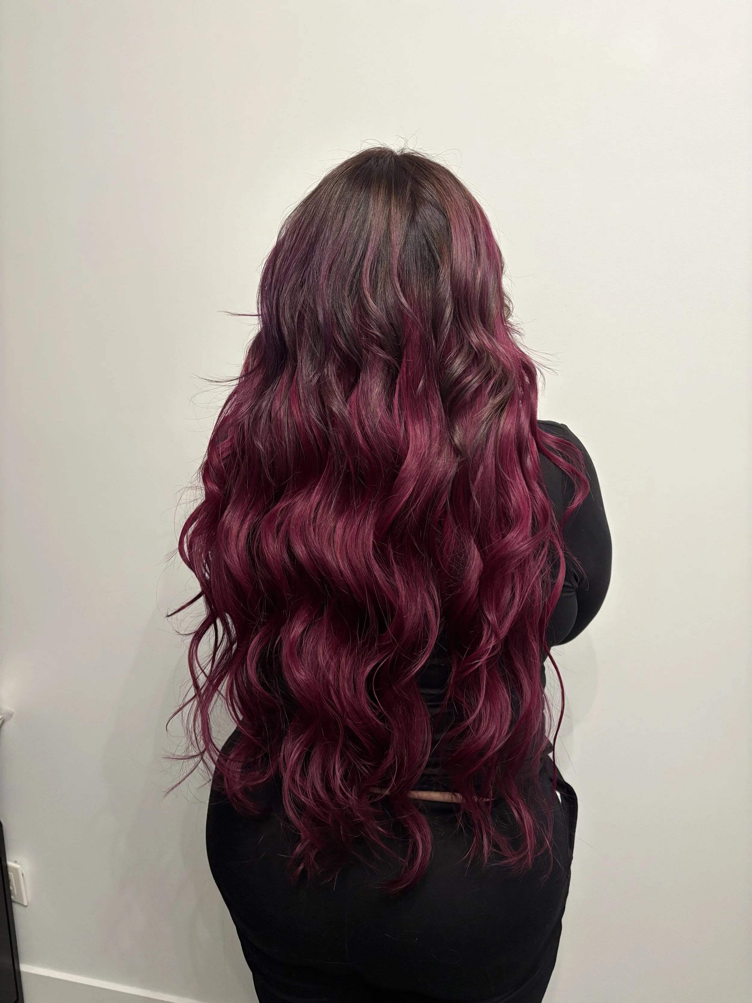 Woman with long, wavy hair in shades of pink and purple, standing against a plain white wall, wearing a black outfit.