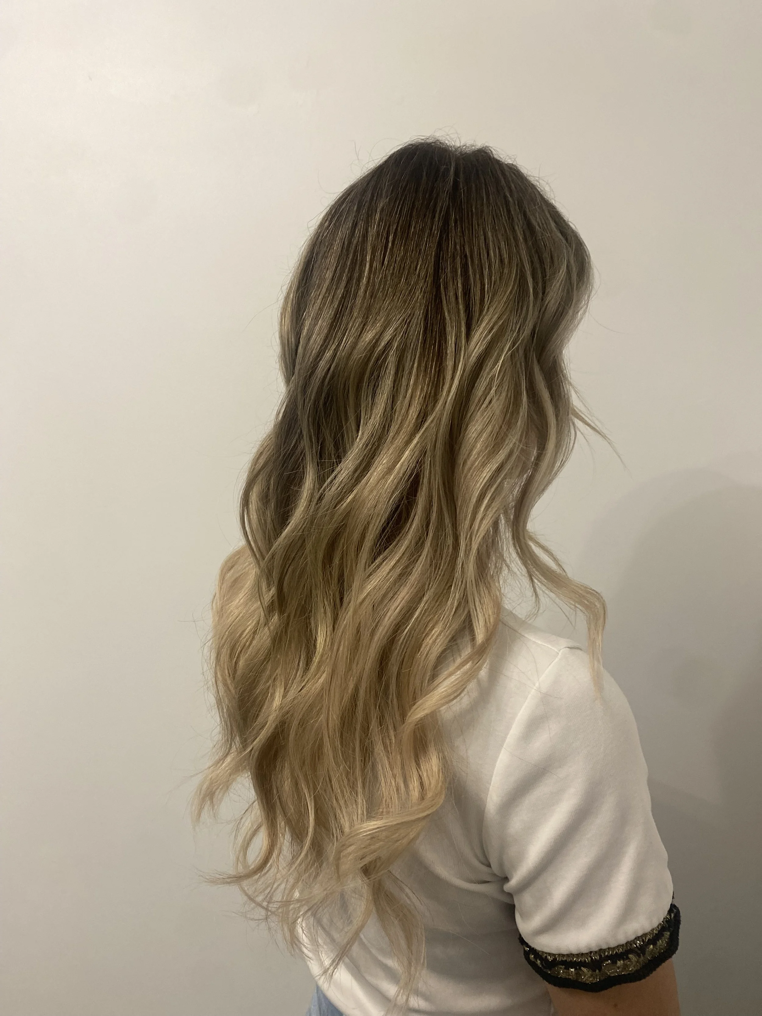 Side view of a woman with long, wavy blonde hair wearing a white shirt with black and gold trim on the sleeves, standing against a plain white wall.