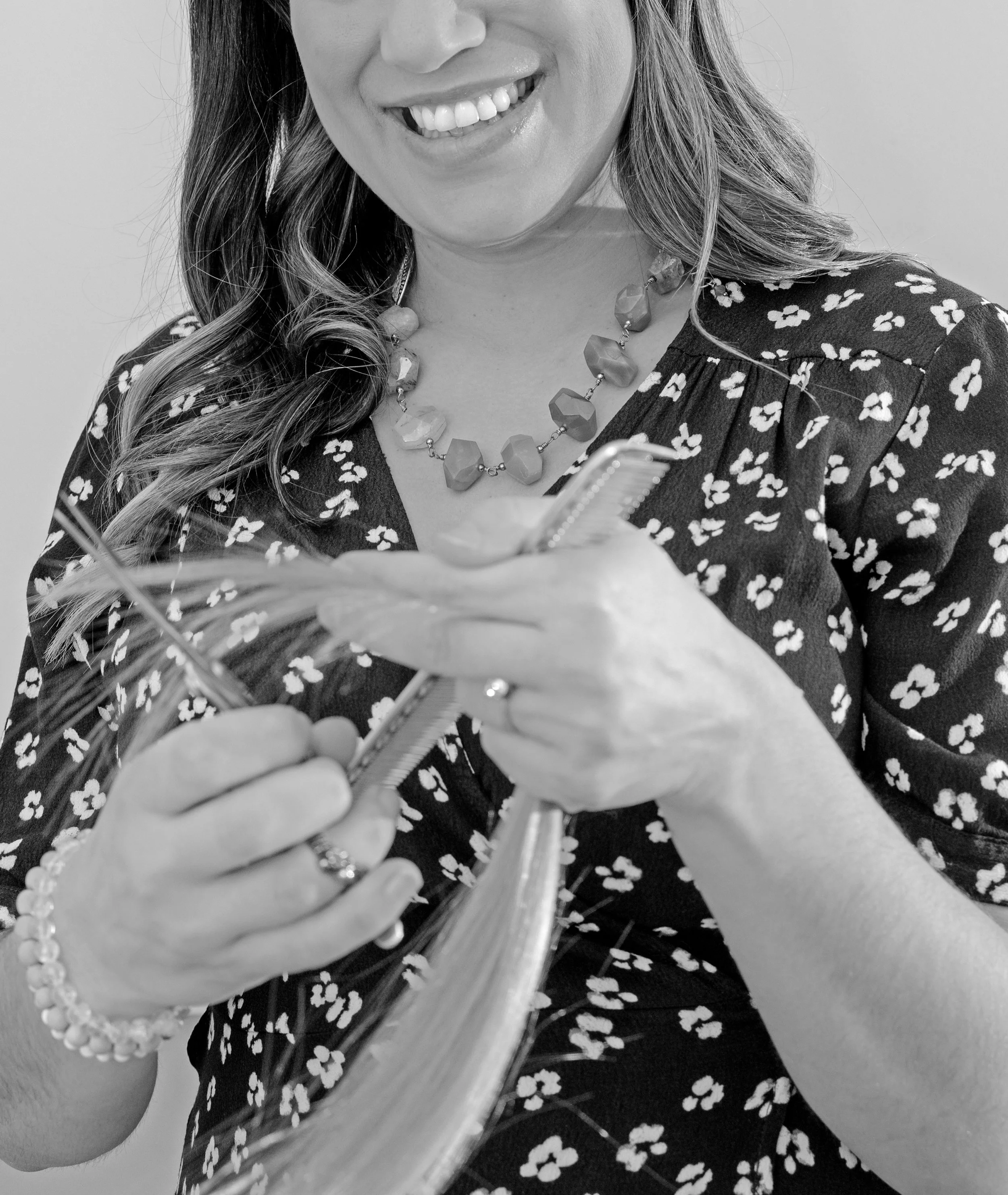 A woman smiling while holding a hairbrush and a feather, wearing a floral dress and a beaded necklace.