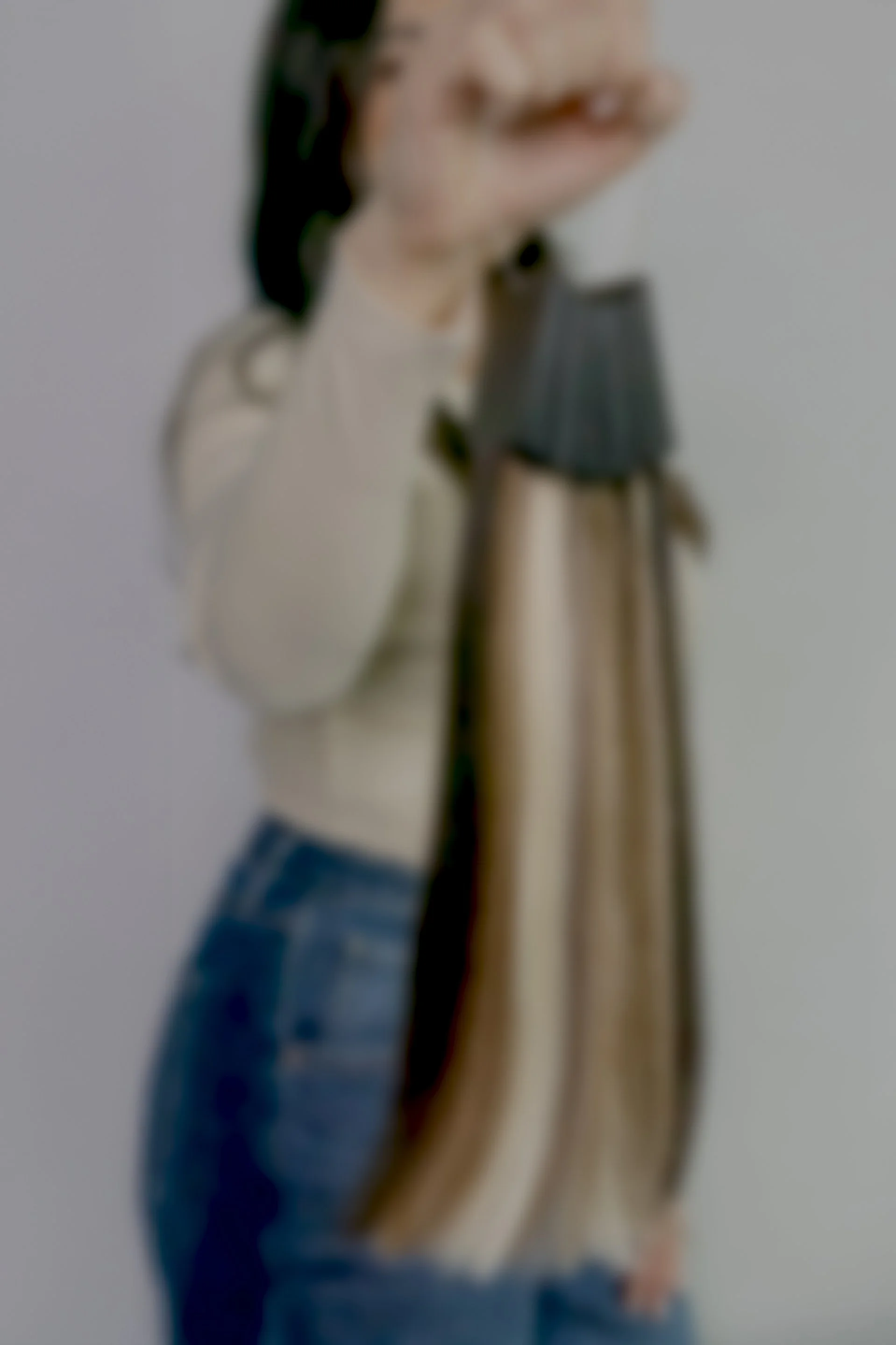 Blurred image of Chelsea holding a hoop of hair extension swatches with her right hand, wearing a beige sweater and blue jeans, against a plain background at the Chelsea Extension Co. Salon in Surrey, BC, Canada.