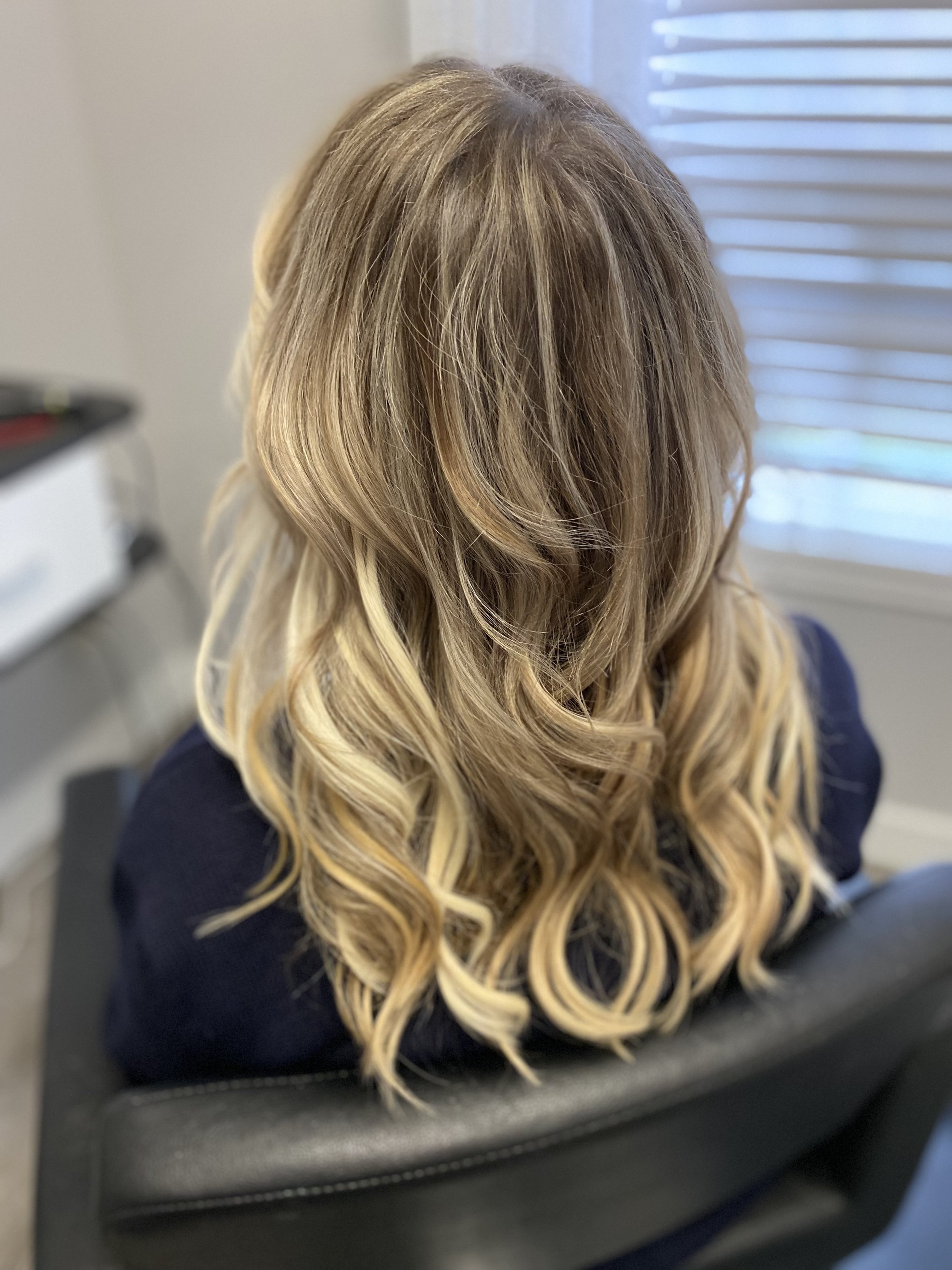 Back view of a person's head with long, wavy blonde hair, sitting in a black chair near a window with blinds.