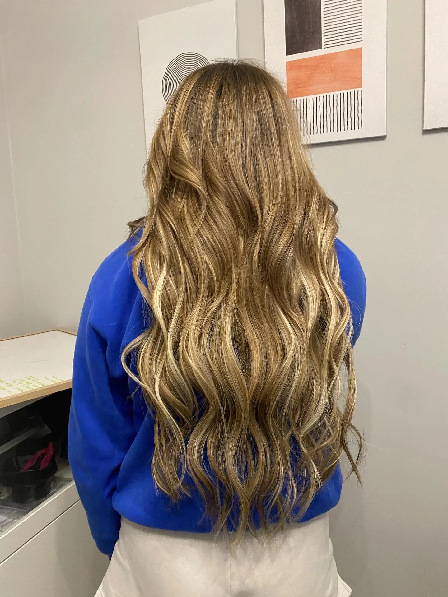 Back view of a woman with long, wavy, blonde hair wearing a blue top, standing indoors near a wall with abstract art.