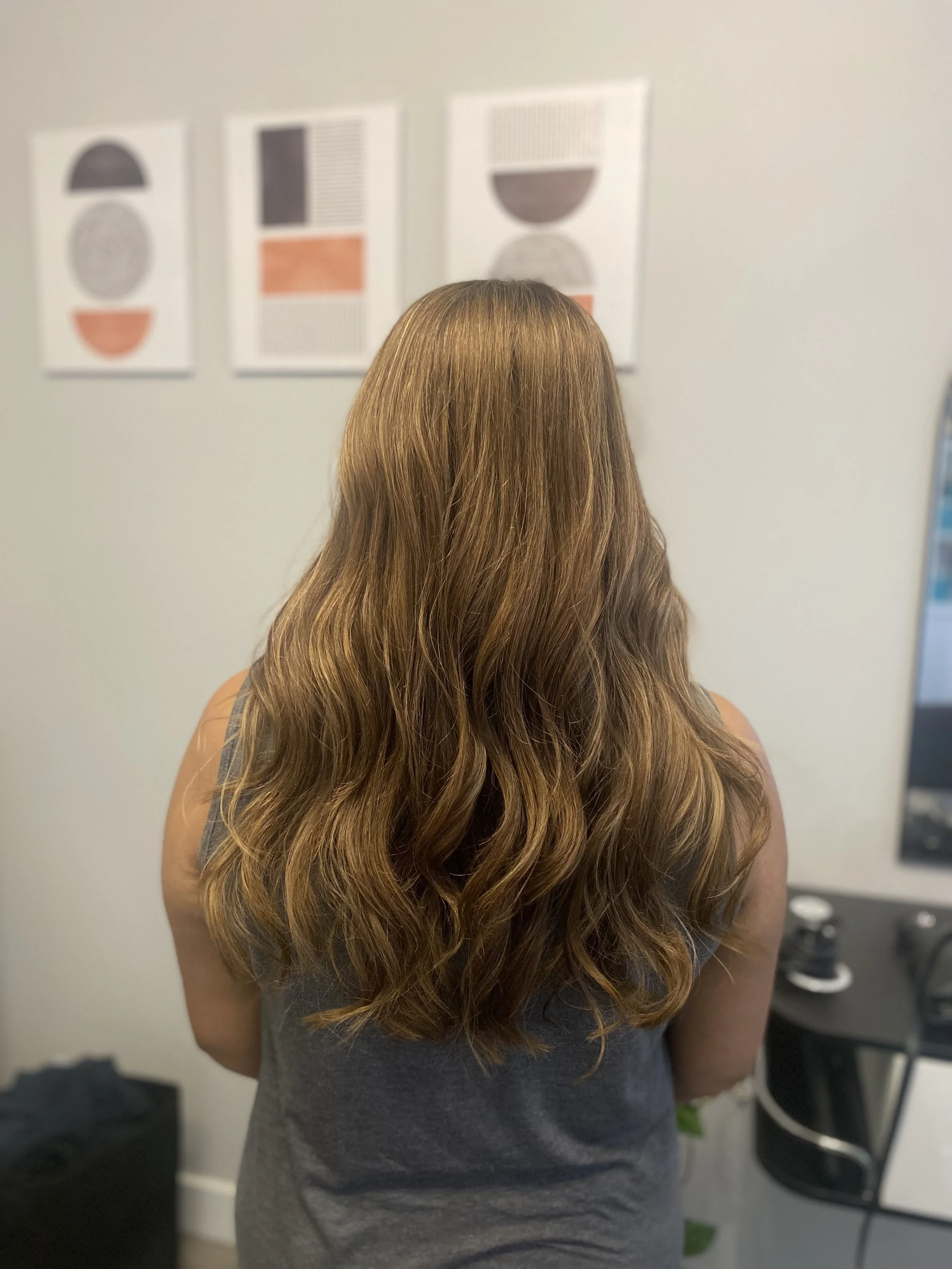 Rear view of a woman with long, wavy, light brown hair standing indoors, looking at abstract art on the wall.