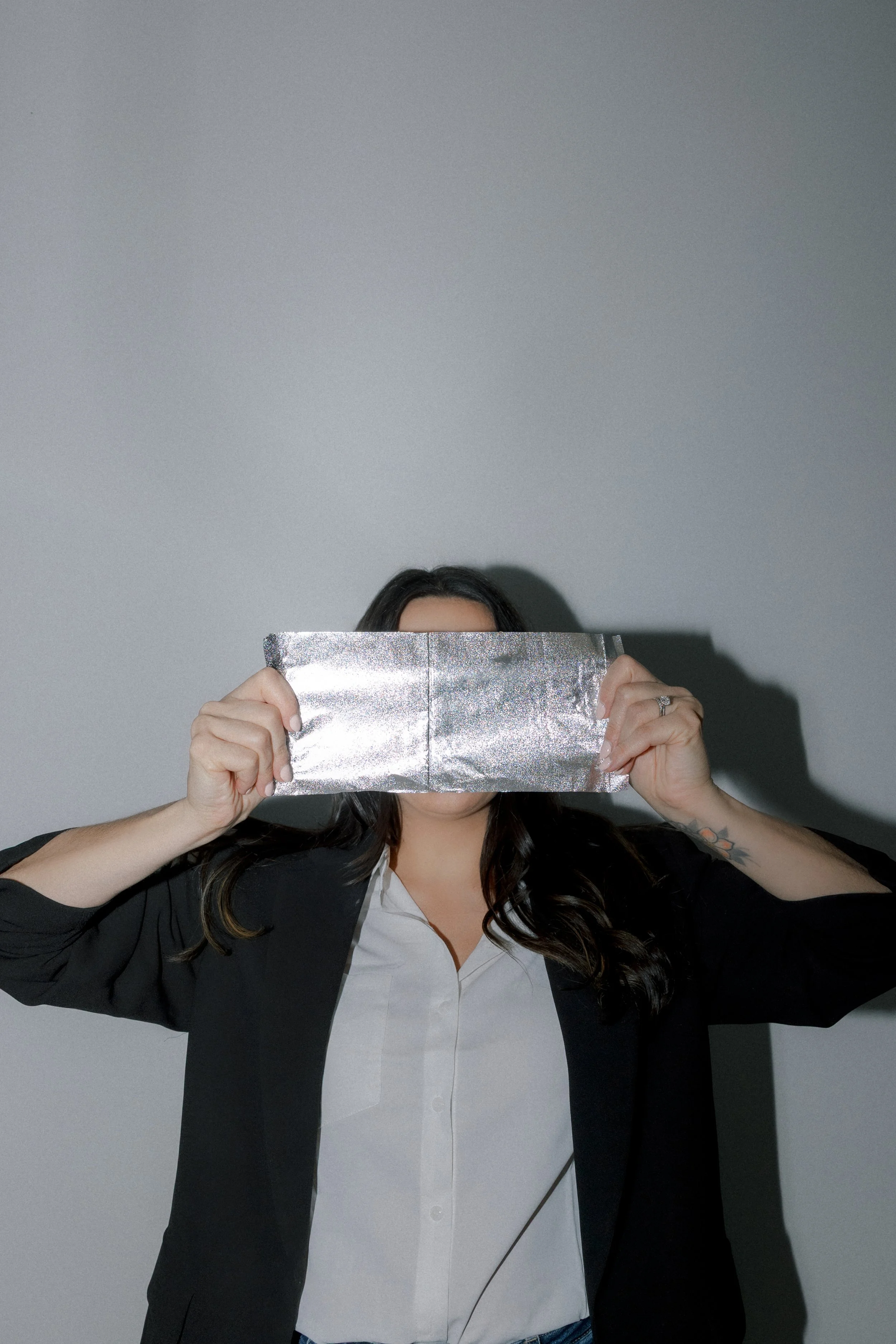 Woman in business attire holding a silver foil object in front of her face, with a plain gray background.