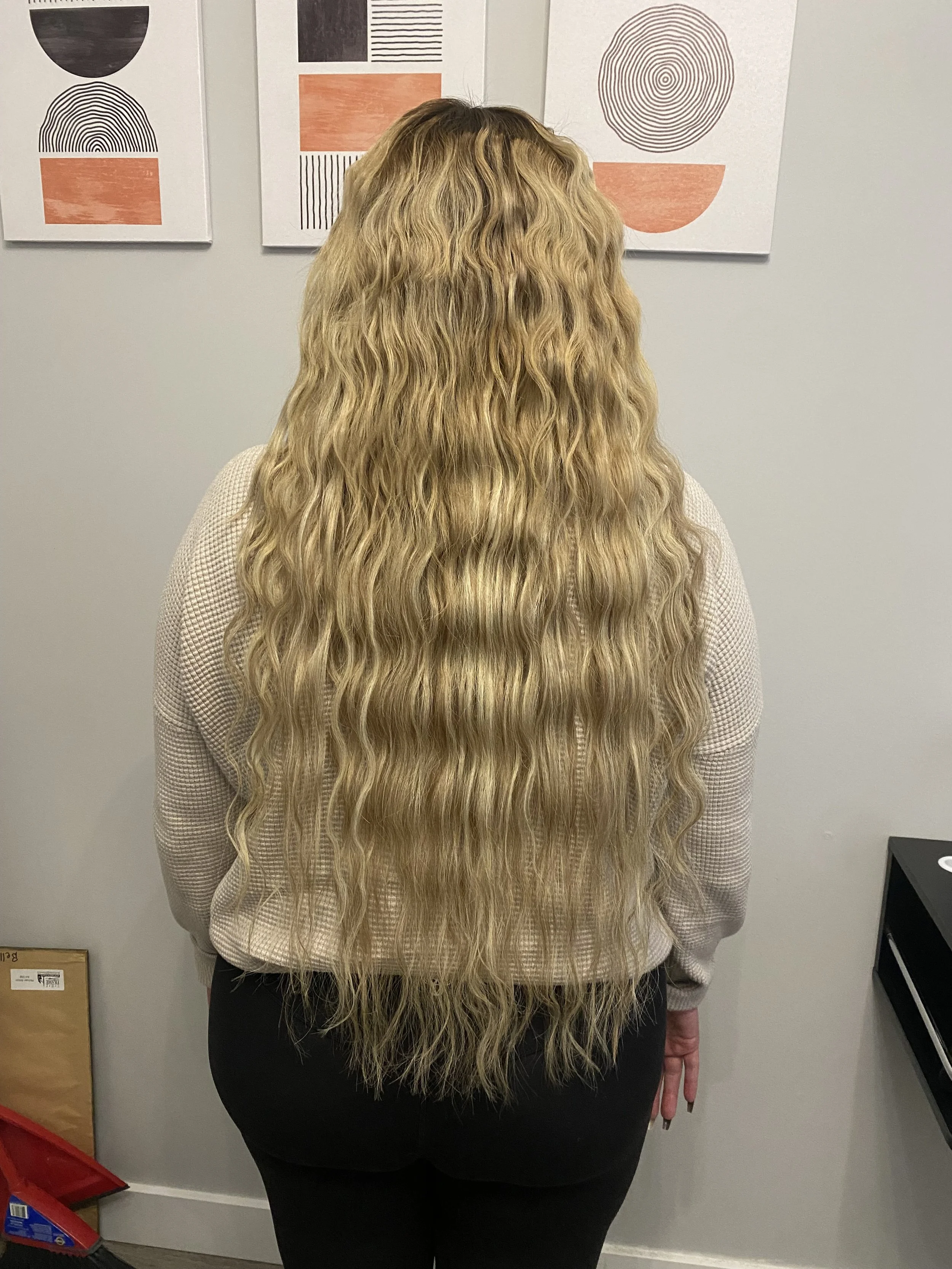 Back view of a woman with long, wavy blonde hair, wearing a beige sweater and black pants, standing in front of wall art.