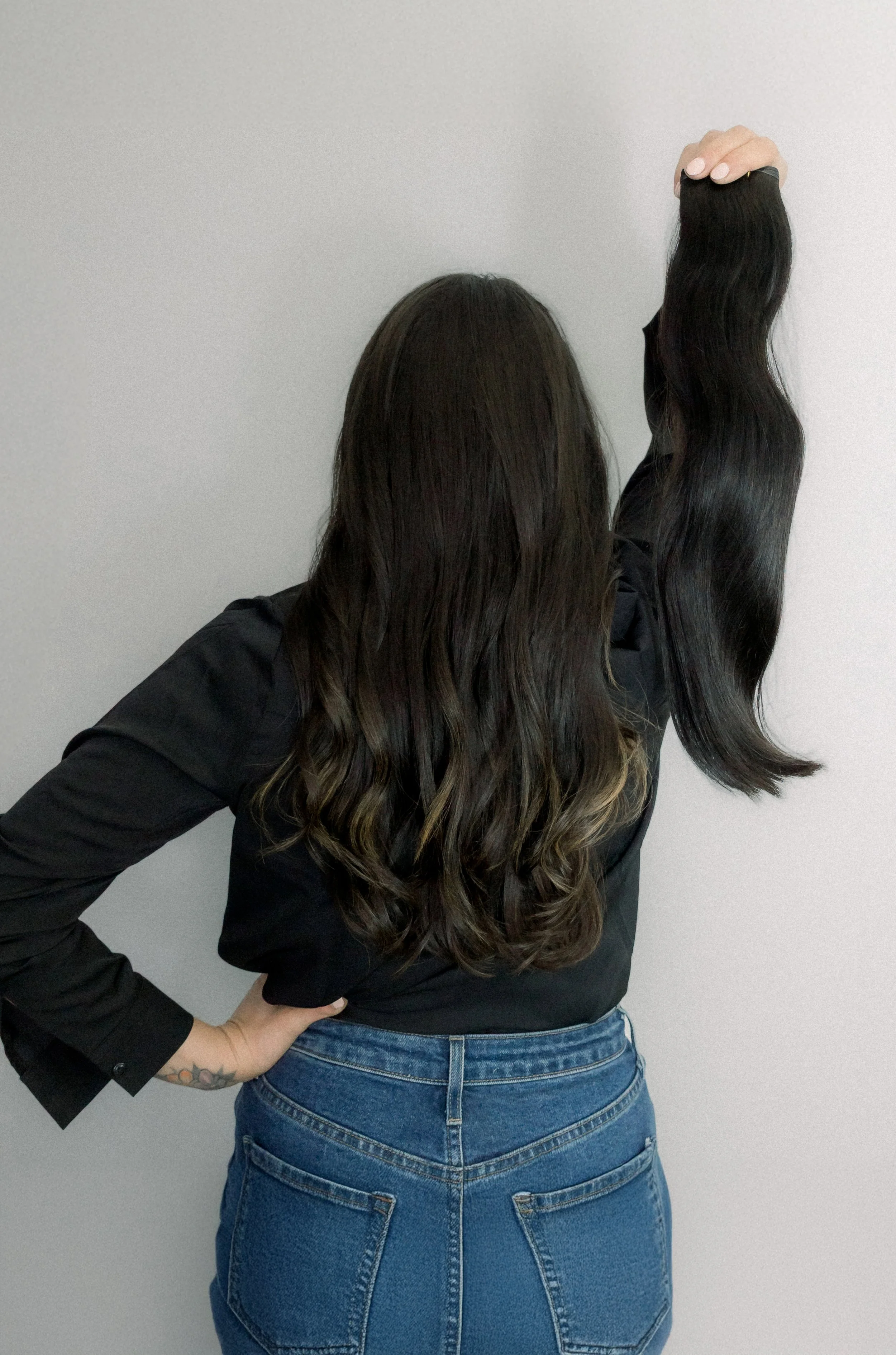 Person with long, dark, wavy hair holding a strand of hair in front of a plain wall.