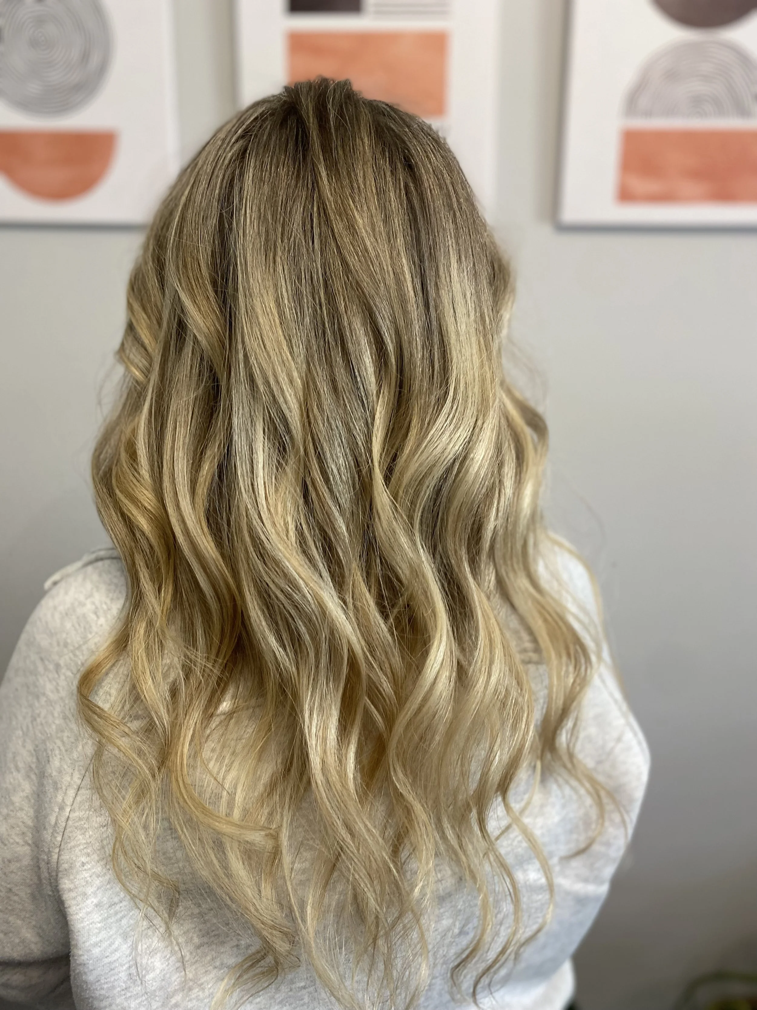 Back view of a woman with long, wavy blonde hair standing indoors against a white wall with abstract orange and black artwork.