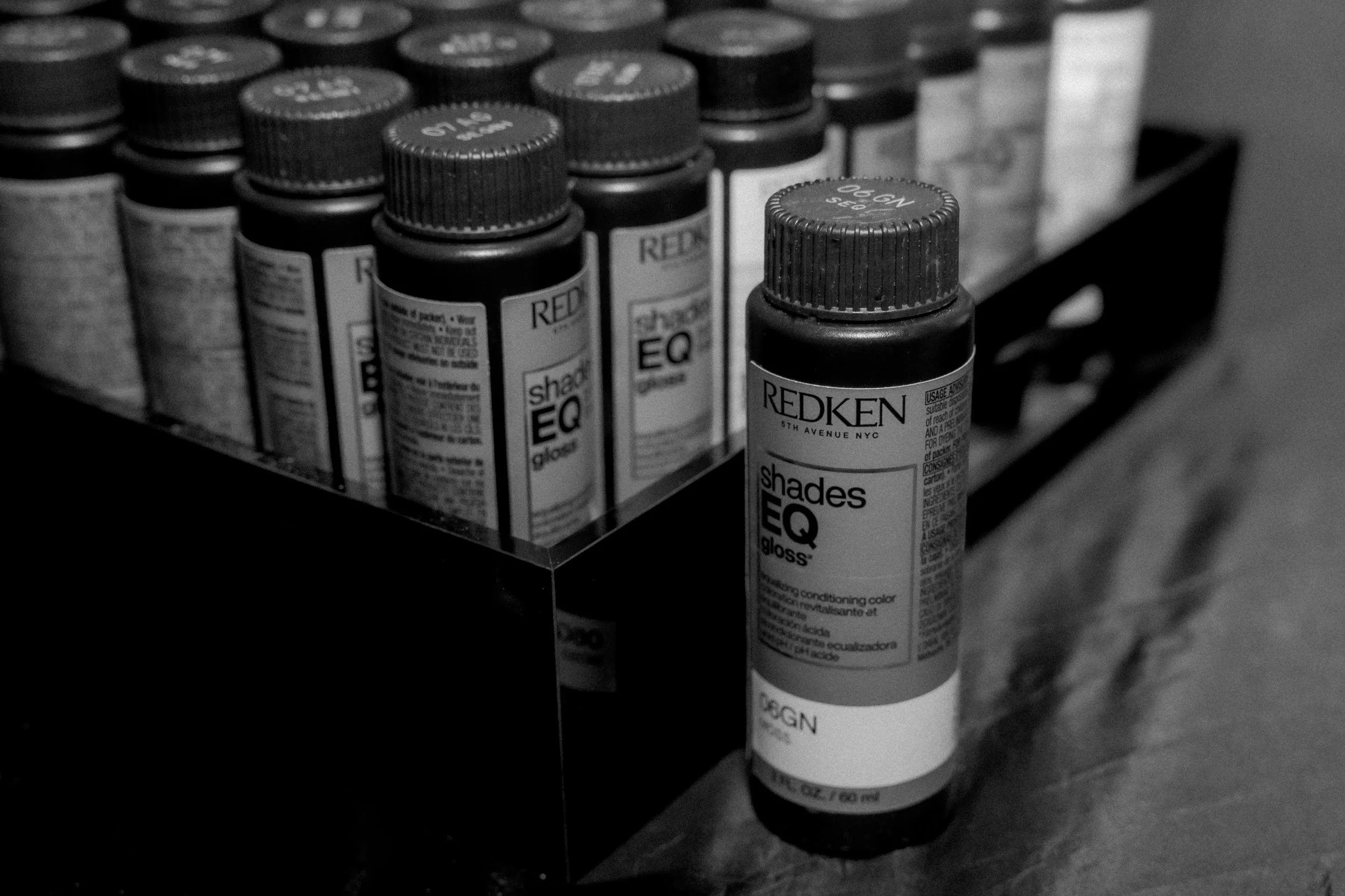 A black and white photo of a collection of Redken Shades EQ gloss hair colour bottles on a black shelf, with one bottle placed outside the shelf container in front at the Chelsea Extension Co. Salon in Surrey BC Canada.