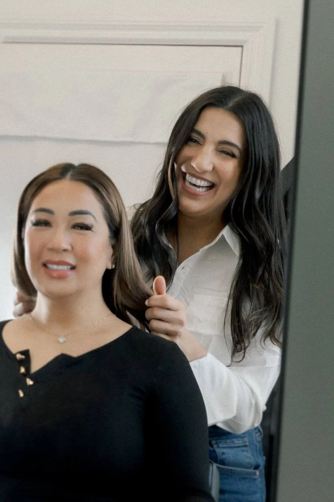 A hairstylist with long dark hair and a white shirt styling the hair of a woman with shoulder-length brown hair wearing a black top, both smiling in front of a mirror.
