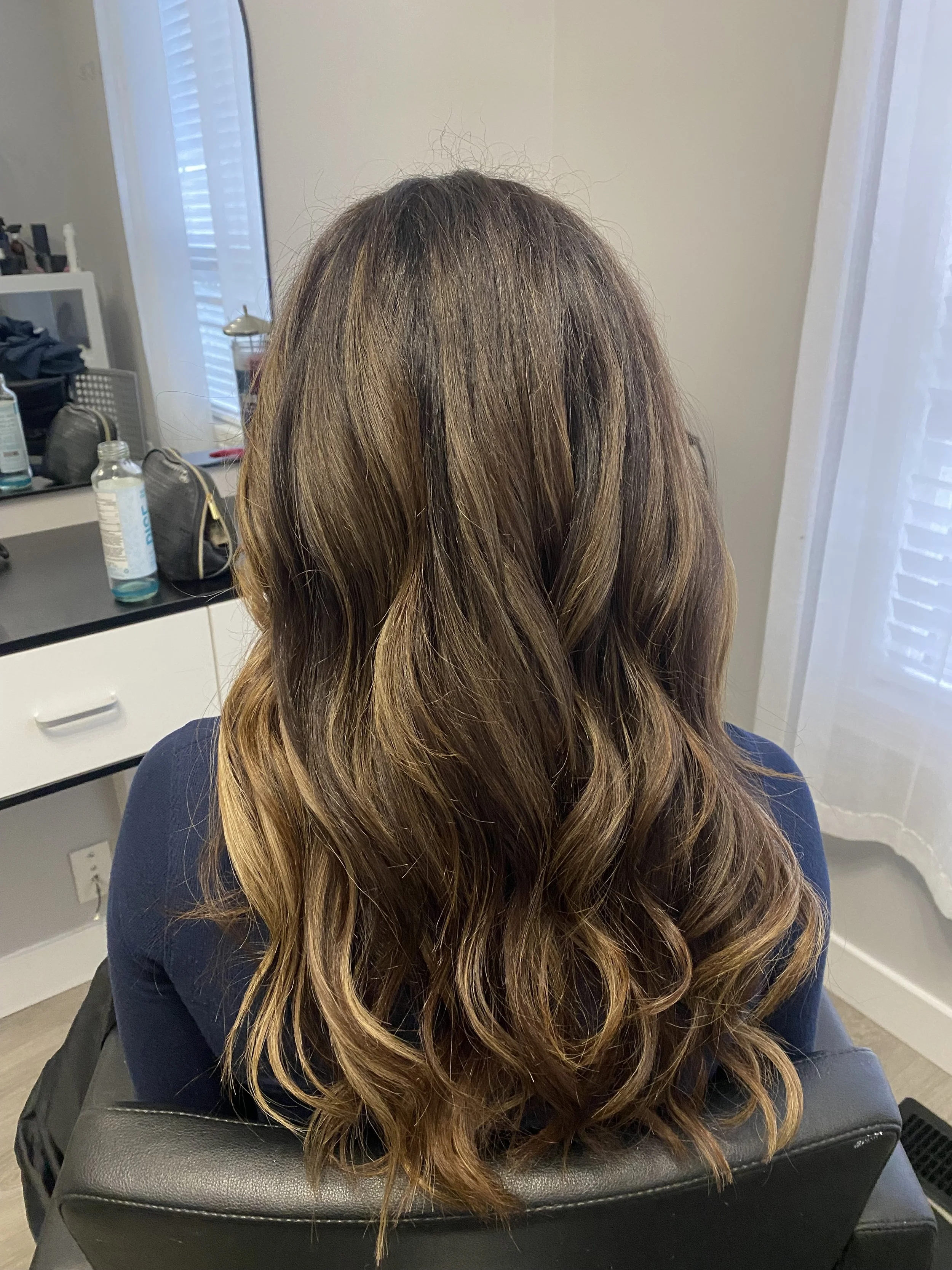 Back of a woman with long, wavy, brown hair with highlights, sitting in a salon chair.