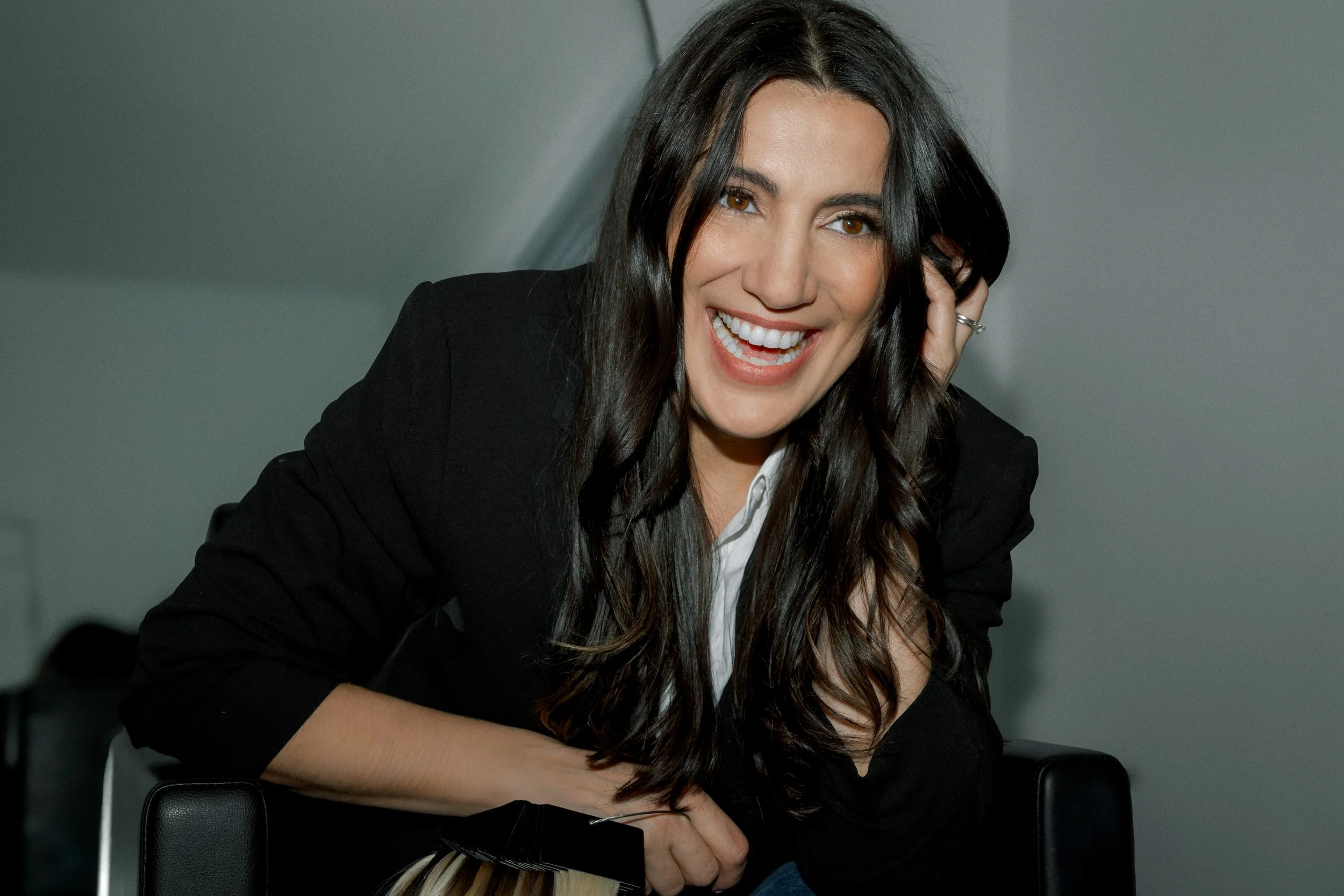 Chelsea, leaning her head with her left hand holding her dark shiny hair, dressed in a black blazer and white shirt, holding a hair extension swatch fan in her right hand, leans over a black chair smiling with teeth, in Surrey, BC, Canada.
